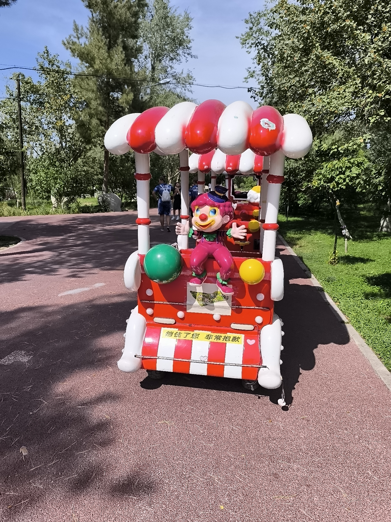A clown-themed children's amusement ride sits on a red paved path outdoors. The ride is designed to resemble a train car with a canopy supported by white poles with red stripes. A colorful clown figure with a wide smile and blue hair is seated in the ride. The clown is wearing a pink jumpsuit with green accents and a black hat. In front of the clown are a green ball and a yellow ball. A yellow sign with Chinese characters is affixed to the front of the ride. The ride is surrounded by greenery, including trees and grass. In the background, several people are walking on a path. The sky is blue and appears clear. The scene is lit by sunlight, casting shadows on the path.