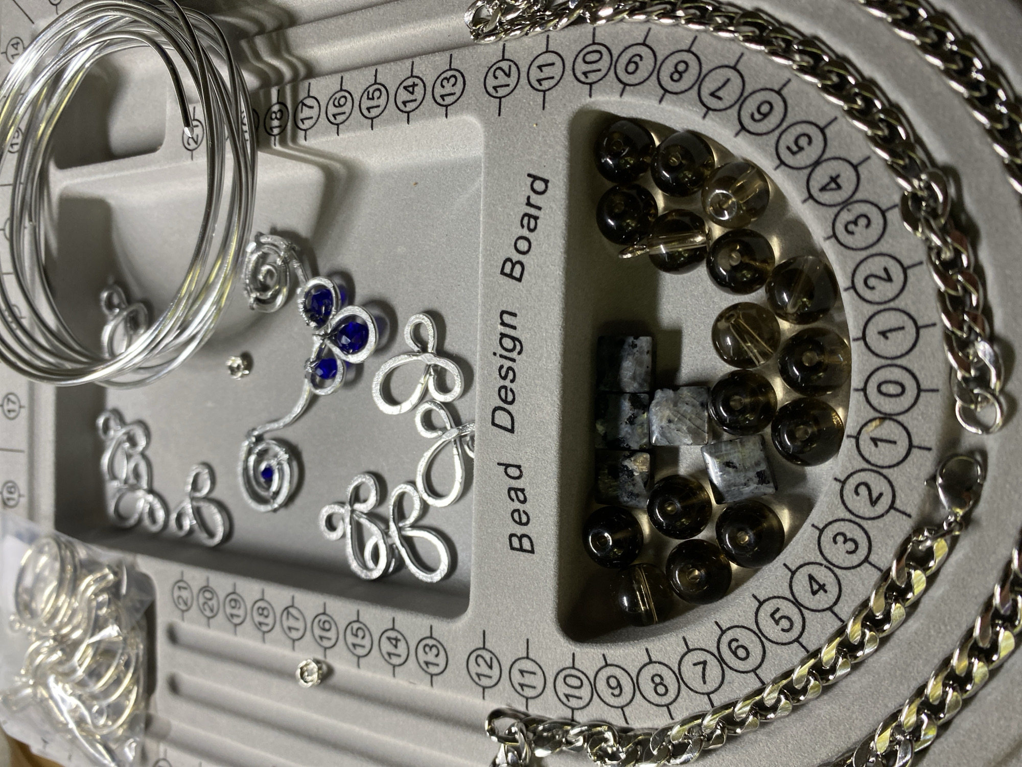 The image shows a close-up, high-angle view of a bead design board. The board is gray and has a textured surface with a grid of numbered circles and lines, indicating measurements in inches. On the board, there are various jewelry-making supplies. In the upper left corner, there is a plastic bag filled with silver-colored jump rings. To the right of the jump rings, there are several silver-colored metal findings with decorative swirls. In the upper right section of the board, a large spool of thin, silver-colored wire is coiled. In the center of the board, arranged in a semi-circle, are several small, clear beads with a smoky brown tint, and a few square beads with a gray, speckled pattern. Two pieces of silver-colored chain are visible in the foreground, one on the left and another on the right, with one end of the right chain featuring a clasp. The background is out of focus. The scene is indoors and appears to be a workspace for crafting jewelry. There are no people visible in the image. The location is not specified in the image or its metadata.