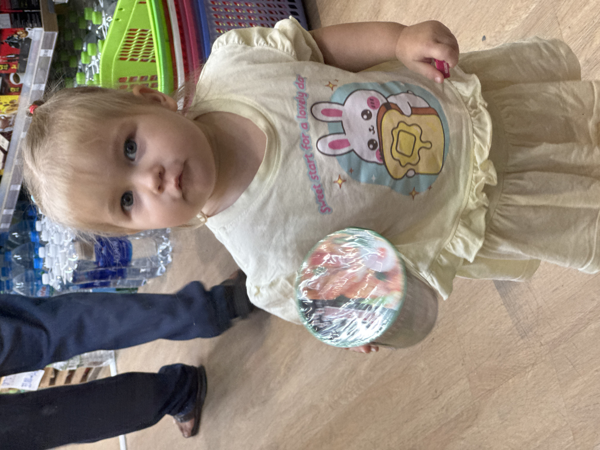A young Caucasian girl, approximately 2-3 years old, stands in a grocery store aisle. She has blonde hair styled in pigtails and blue eyes, and is wearing a light yellow t-shirt with a graphic of a bunny holding toast and the words "Sweet start for a lovely day." She is holding a cylindrical object wrapped in clear plastic with a colorful, blurry pattern. Behind her, to the right, are stacked green, red, and blue plastic baskets. Further in the background, shelves are stocked with goods, and bottles of water are visible. To the left of the girl, the legs of an adult wearing dark pants and sandals are visible, suggesting they are accompanying her. The flooring appears to be wood laminate. The environment suggests a retail setting typical of a supermarket or convenience store, which could be found anywhere in Nha Trang, Vietnam.