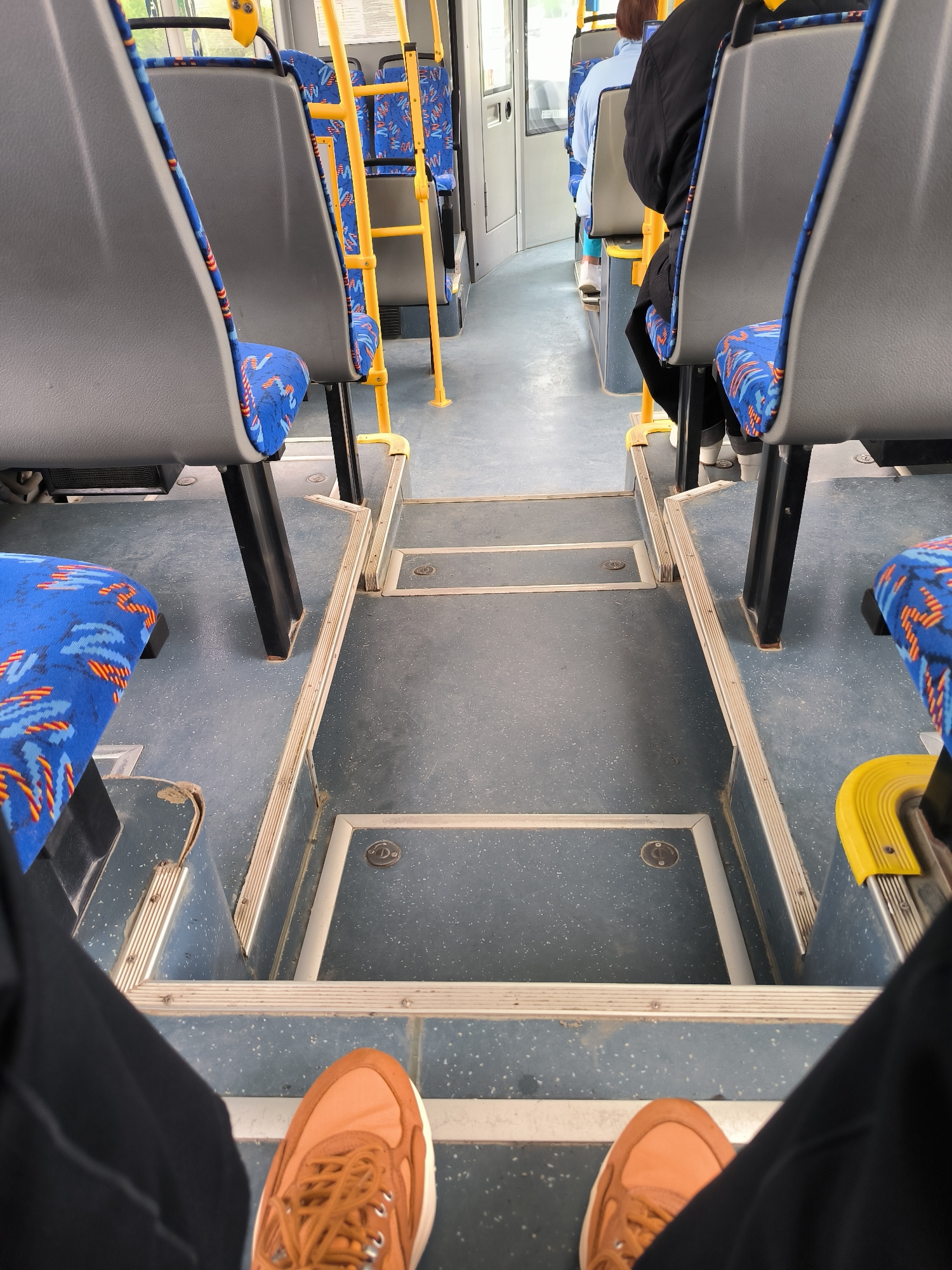 The image shows the interior of a public bus, likely in Yuzhno-Sakhalinsk, Russia, looking towards the front from a seated position. The perspective is low, focusing on the floor and the lower parts of the seats. Two seats with grey backs and blue patterned cushions are visible in the foreground and to the left. The floor is a speckled dark blue material with several steps and raised edges. Yellow grab bars are present along the aisle. In the background, other passengers are seated, including one person in a light blue shirt and another in a dark jacket. The lighting suggests it is daytime. The overall environment is utilitarian and characteristic of public transportation.