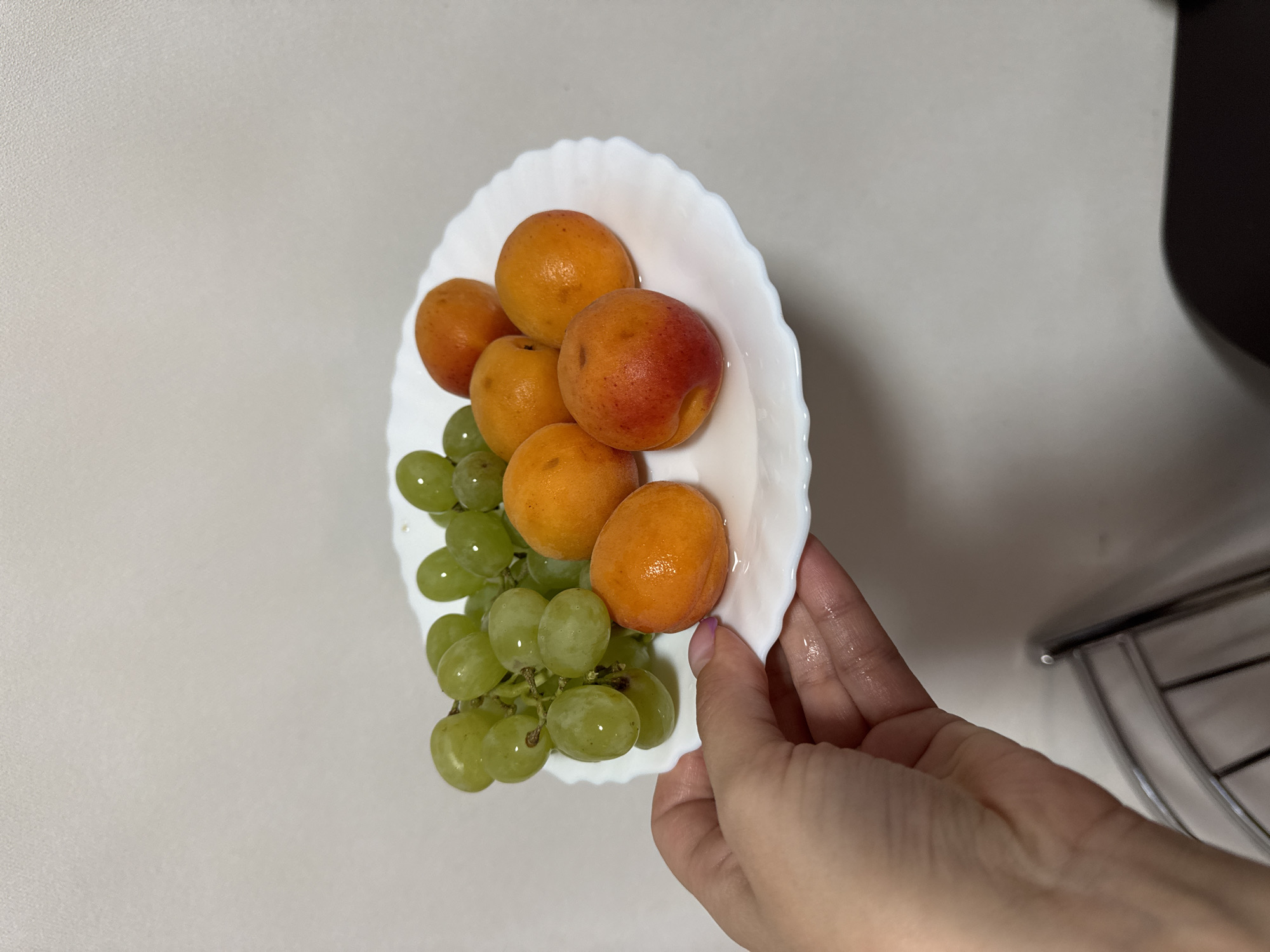 A white plate with a bunch of green grapes and six apricots is held by a person's hand. The plate is positioned in front of a light grey countertop. To the right of the plate is a blurred metal structure, potentially part of the kitchen environment. The scene appears to be indoors. There is no indication of a specific location in Chaikovsky, Russia.