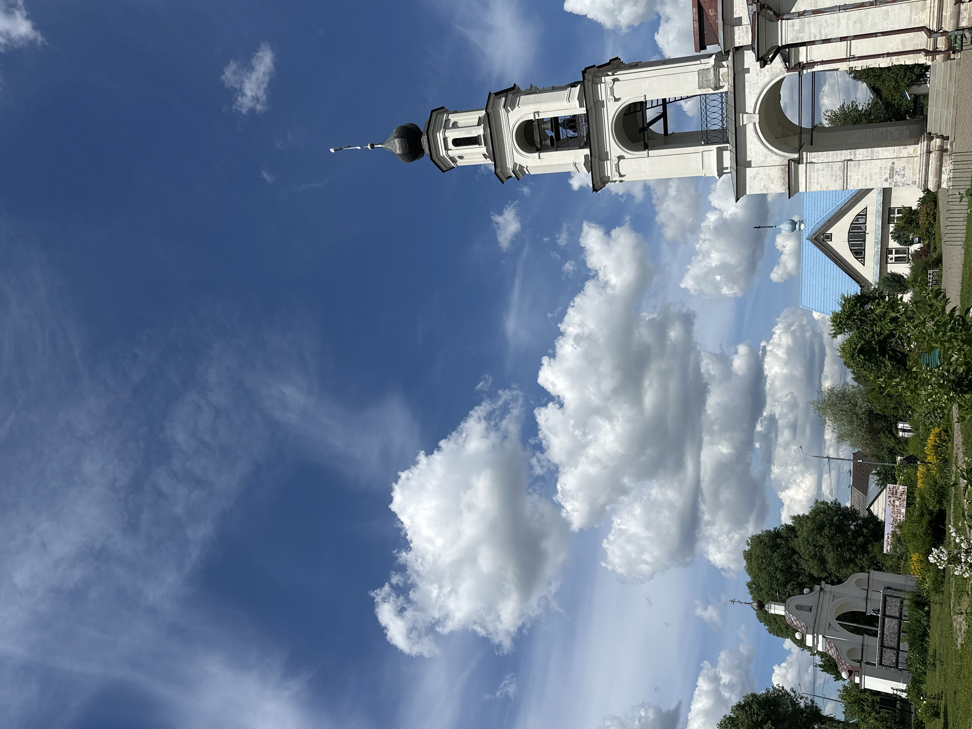 In the city of Erino, Russia, a bright, partly cloudy sky forms the backdrop for a scene dominated by religious architecture. A tall, white bell tower with a dark, onion-shaped dome stands prominently on the right, featuring arched openings and a cross at its apex. To the left, a smaller, white arched structure with a cross also suggests a religious site. In the mid-ground, a building with a bright blue roof and a smaller dome is visible, surrounded by lush green trees. The overall environment appears to be a park or public space, with green grass and blooming yellow flowers in the foreground. The lighting suggests it is daytime.