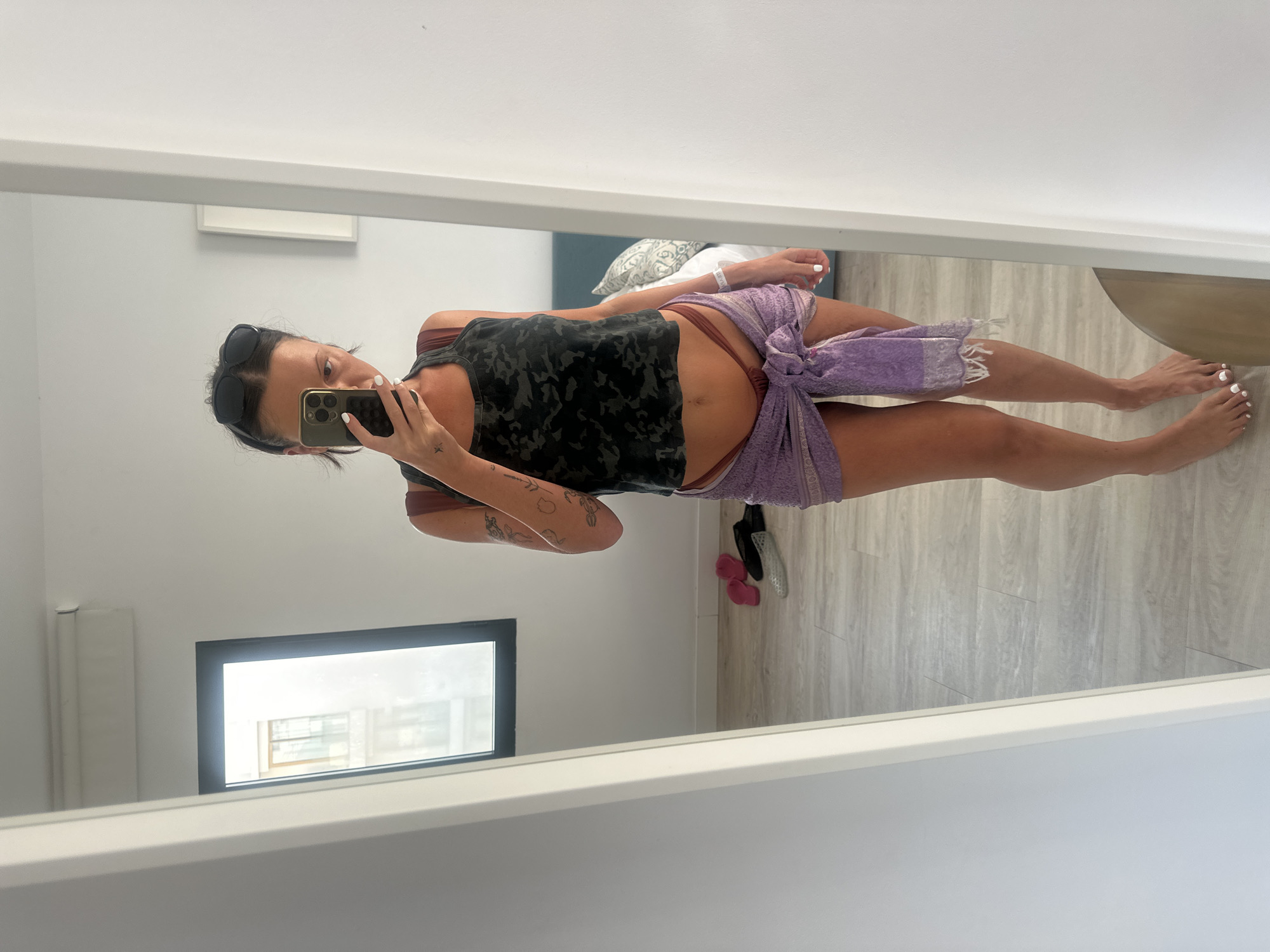A person is captured taking a mirror selfie in a well-lit indoor setting, likely a room in Eivissa, Spain. The image is taken from a slightly elevated perspective, reflecting the person's full body.
The central figure, appearing to be female, is holding a modern smartphone to take the picture, partially obscuring their face. Sunglasses are pushed up onto their head. They are wearing a dark, camouflage-patterned tank top and dark reddish-brown high-cut briefs, with a light purple, patterned sarong tied loosely around their waist. Their fingernails and toenails are painted white, and a small tattoo is visible on their inner left forearm. A white wristband, possibly from an event or medical facility, is worn on their right wrist.
The background, visible in the mirror's reflection, portrays a modern and minimalist room. White walls and light wooden floorboards dominate the space. Behind the person, a bed with white and patterned pillows can be seen. To the left, a window reflects glimpses of other buildings, indicating an urban or built-up area. The bright natural light streaming into the room suggests it is daytime. The room appears clean and tidy, potentially a hotel room or contemporary apartment.