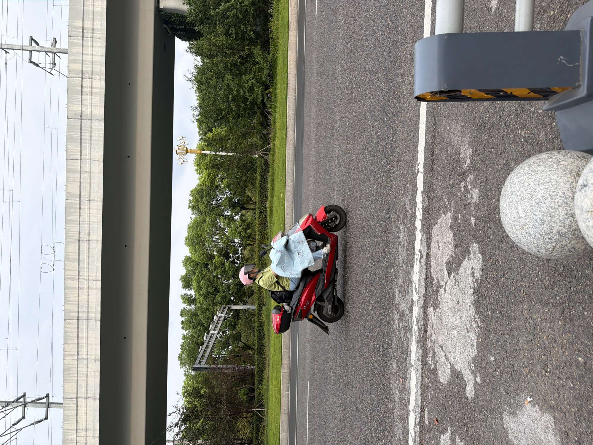 The image shows a person riding a red electric scooter on a paved road, possibly in Jiangjin, China. The person is wearing a pink helmet and a light green shirt, and is covered by a light blue blanket. They are riding under an elevated overpass or bridge. In the background, there are trees and a streetlight. The road is asphalt and appears to be a multi-lane road. There are white lines marking the lanes. In the foreground, there are some concrete bollards and a grey metal barrier. The sky is overcast. The scene appears to be a typical urban or suburban environment.