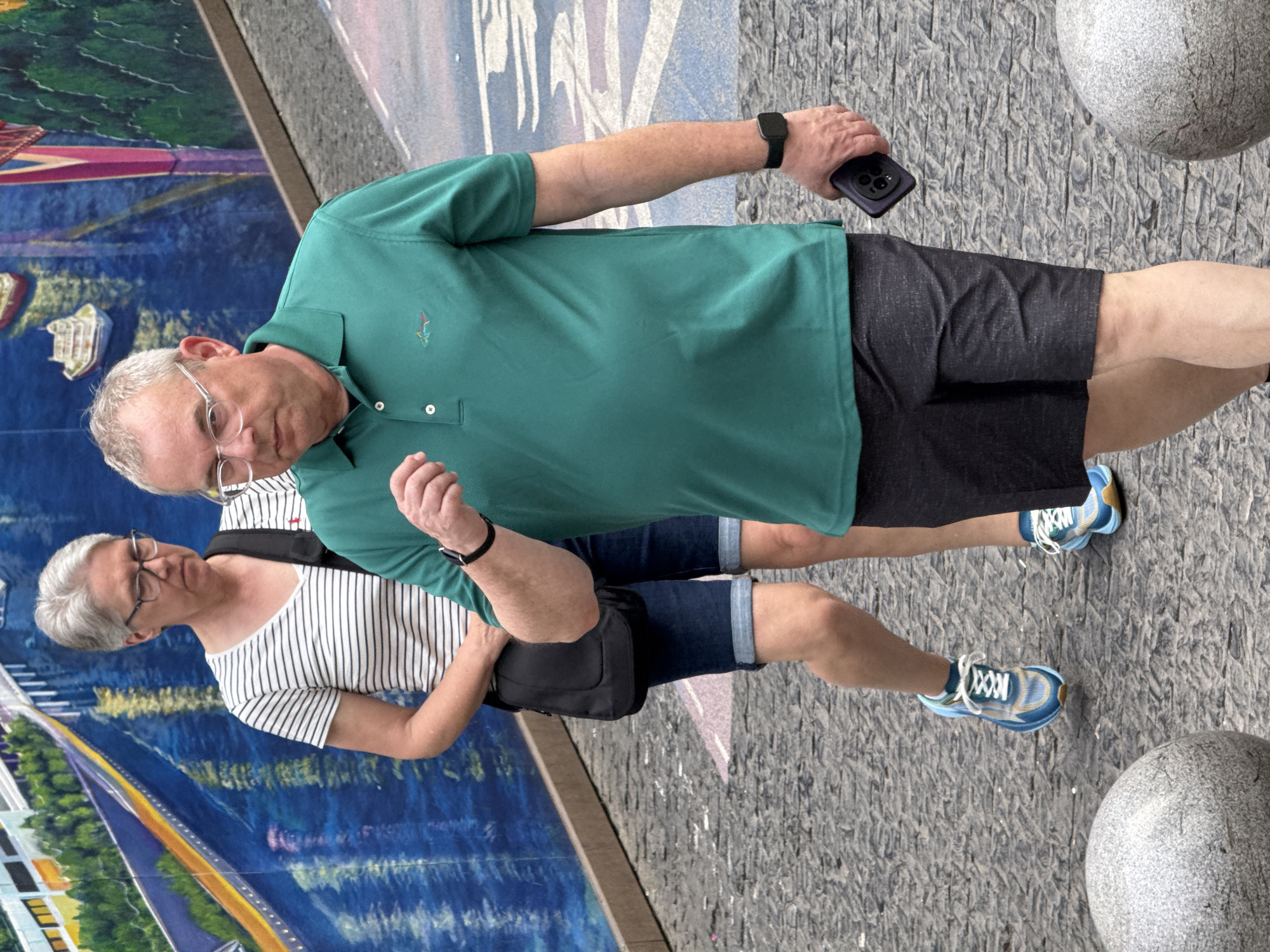 Two people stand on a textured gray surface beside a colorful mural. The person on the left wears a white and black striped shirt, blue denim shorts, and black framed glasses. A black bag hangs over their shoulder. The person on the right wears a green polo shirt, black shorts, a black wristwatch, and holds a black phone. Both are wearing blue and white sneakers. They are standing near two large gray spheres. The colorful mural features buildings, roads, and bodies of water. The location is Yuzhong District, China.