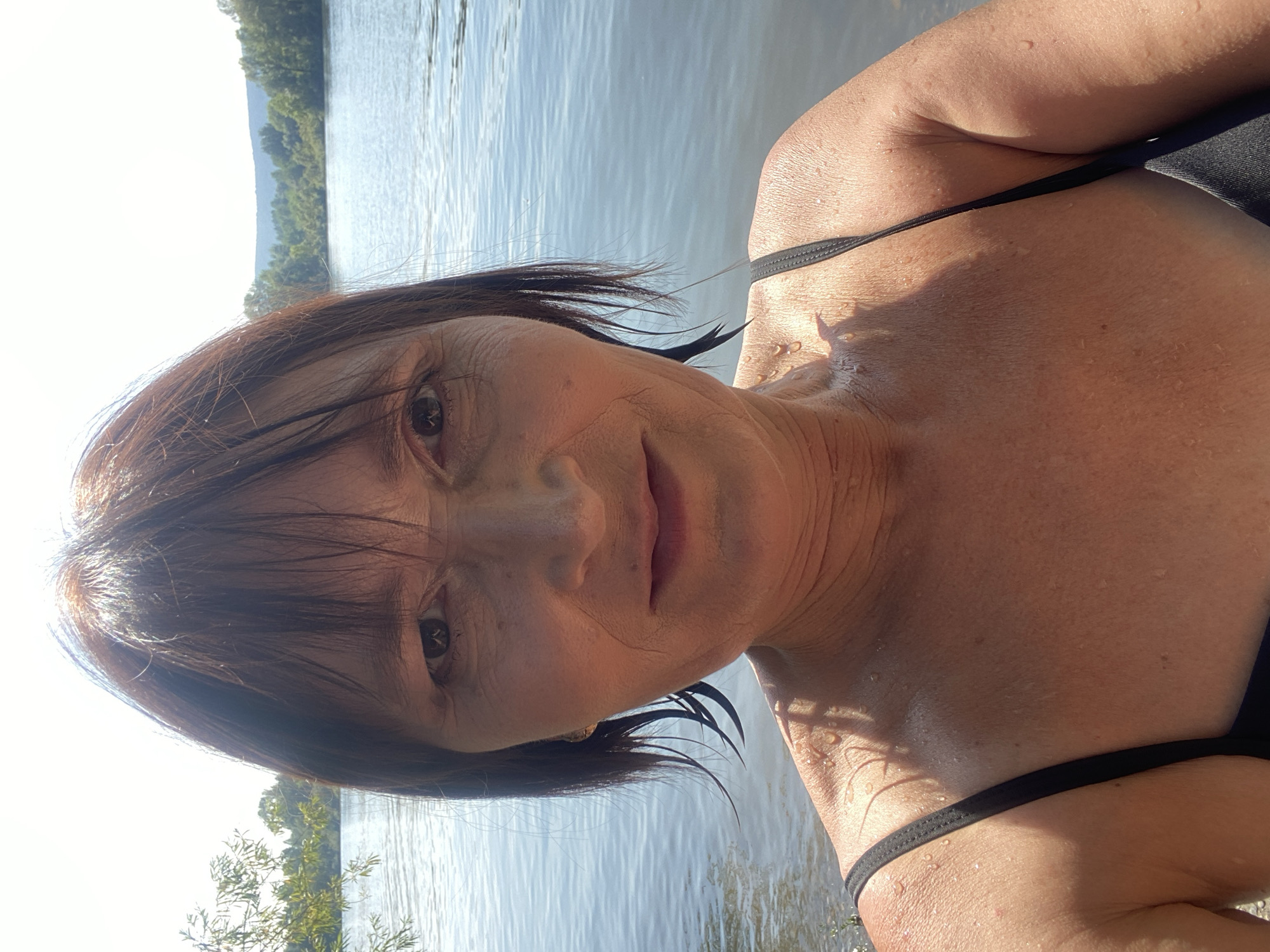 The image shows a close-up, selfie-style shot of a middle-aged woman with shoulder-length, dark brown hair and bangs. She is wearing a black bathing suit. Her face is partially wet, with water droplets visible on her skin and hair. She is looking directly at the camera with a neutral expression. The background consists of a body of water, likely a river or lake, with trees and a hazy sky visible in the distance. The lighting suggests it is daytime, with sunlight illuminating the scene. The location of Novokuznetsk, Russia is not discernible from the image itself.