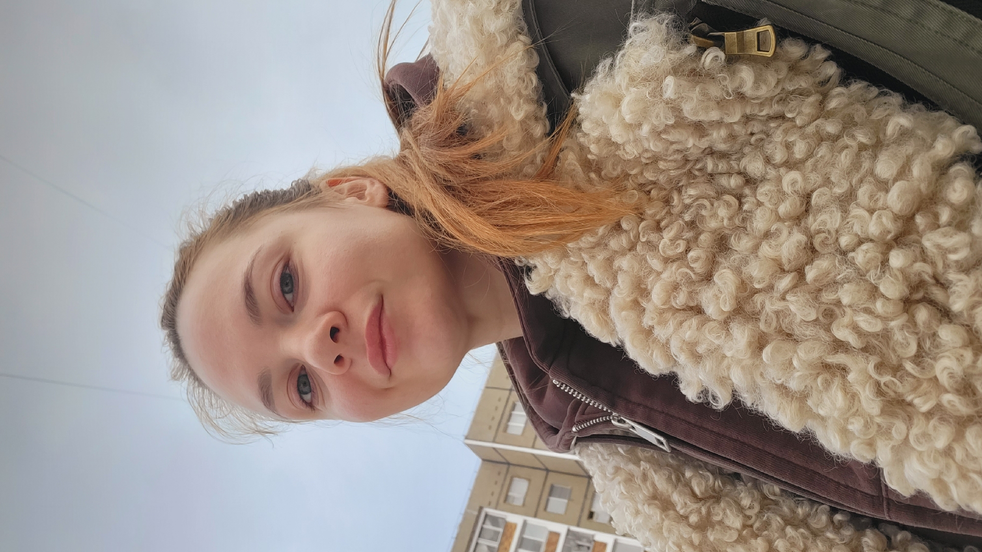 A selfie, rotated counter-clockwise, depicts a fair-skinned woman with light-blue eyes and reddish-blonde hair, possibly in Chaikovsky, Russia, based on the visible building. She is wearing a brown jacket with a thick, cream-colored, sheepskin-like lining around the collar. A multi-story building with windows is visible in the background. The sky appears overcast.