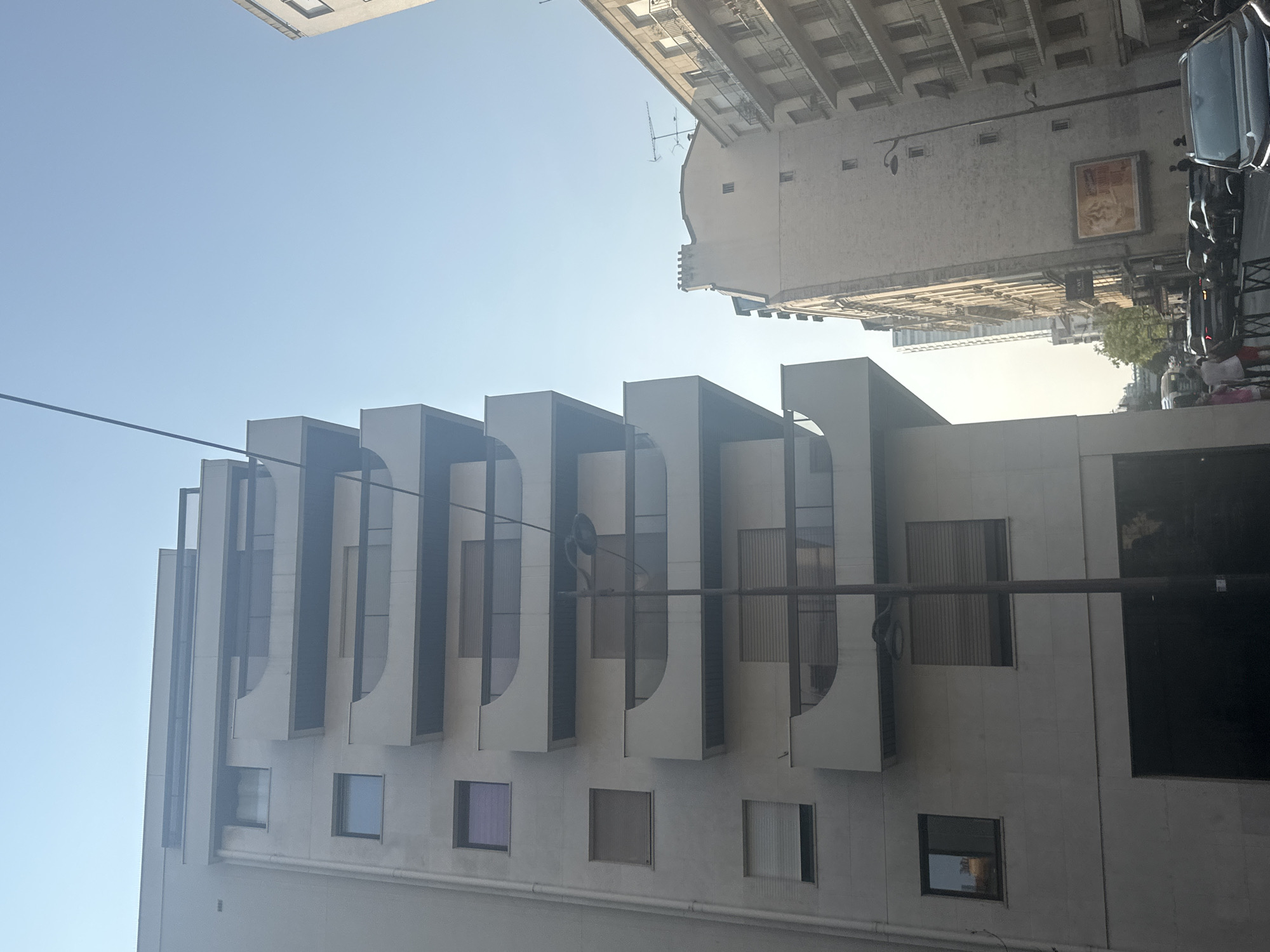 The image depicts a view of Parisian city streets and buildings under a clear sky. The foreground features a modern, light-colored apartment building with balconies on multiple levels. To the right, an older, multi-story building with cream-colored facades and balconies is visible, adorned with antennas on its roof. Further down the street, other buildings of varying heights and architectural styles are present. Several cars, including SUVs and sedans, are parked and driving on the street. A few pedestrians are also visible on the sidewalk. The scene suggests a daytime setting in a dense urban environment.
