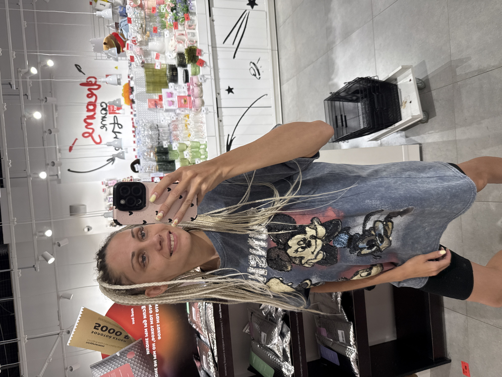 A young woman with blonde braided hair takes a selfie in a brightly lit store. She is wearing a faded blue t-shirt with a graphic of Mickey Mouse. She is smiling and holding a pink phone with heart-shaped accents. In the background, shelves are stocked with various items, including decorative objects and what appear to be kitchenware. Red and black lettering on a white wall is partially visible, with some letters in Cyrillic. A shopping cart with a black wire basket is on wheels to the right. A yellow coupon with the number "2000" in large print is visible in the lower left corner of the image, partially obscuring some text. The overall impression is that of a casual shopping trip. The location context provided is Arkhangelsk, Russia.
