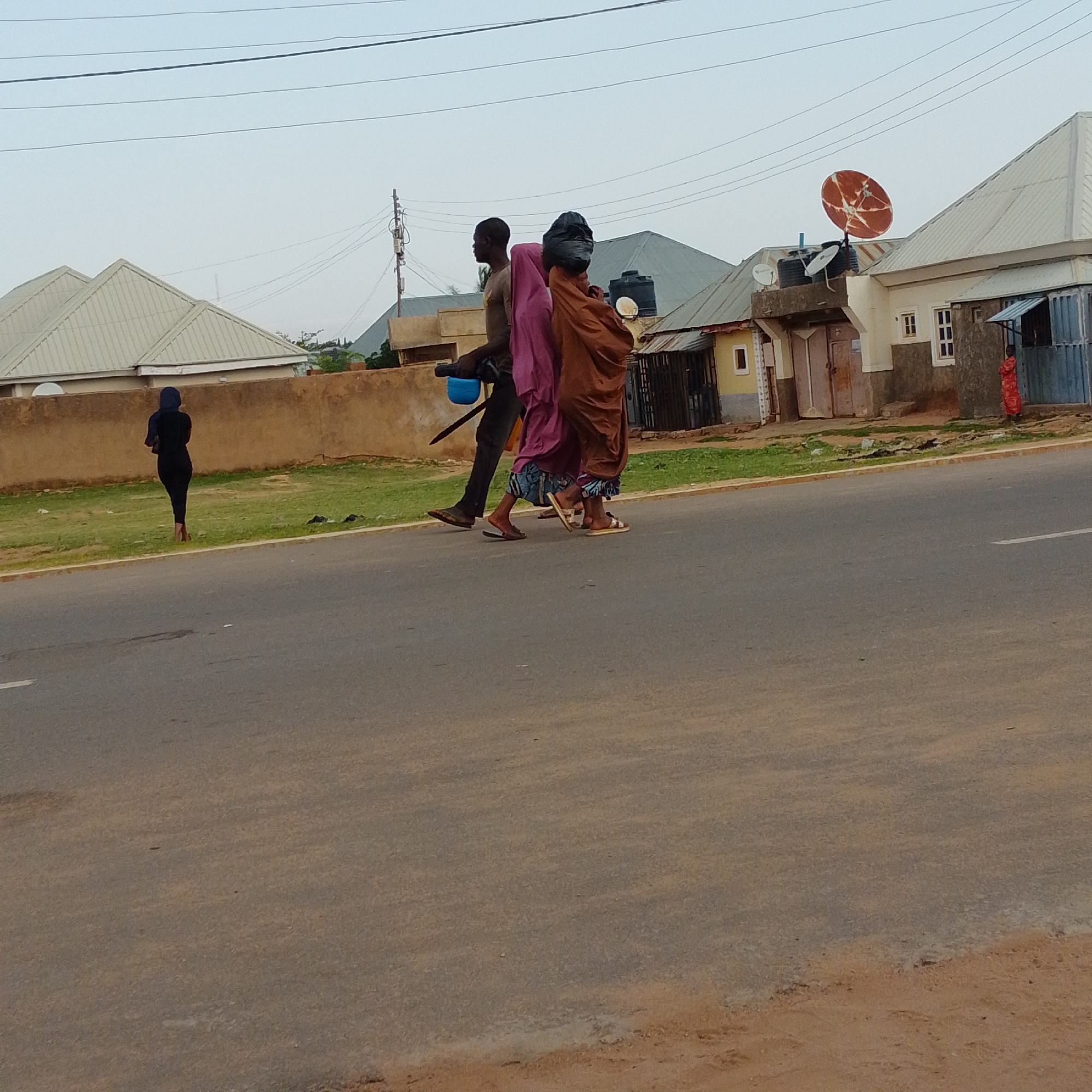 In the city of Gombe, Nigeria, three individuals are walking along a paved road. Two of them, a man and a woman, are walking side-by-side. The man, wearing a t-shirt and dark trousers, is carrying a blue container in his left hand and what appears to be a stick or tool in his right. He is wearing dark shoes. The woman beside him is wearing a long, brown garment and a pink head covering, with a dark head covering on top. She is wearing sandals. A third person, a woman in a black dress and head covering, is walking alone in the distance on the grassy shoulder of the road, facing away from the viewer. In the background, residential buildings with peaked roofs are visible, along with a satellite dish mounted on top of one of the structures. The sky is overcast.