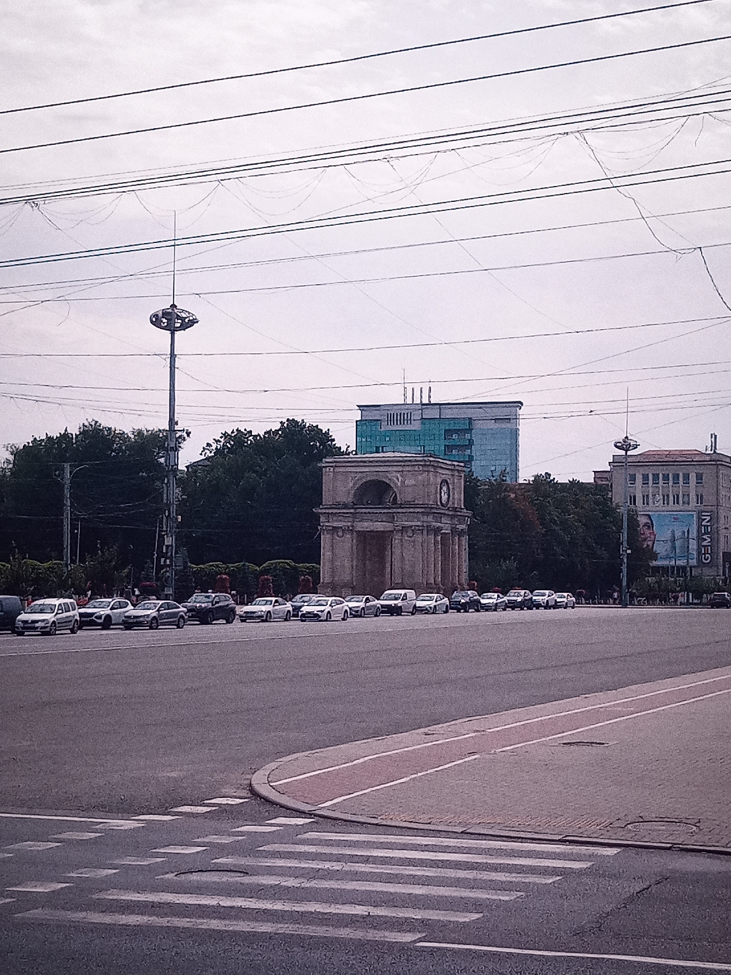 The image captures a street scene in Chișinău, Moldova, featuring the Triumphal Arch and the Great National Assembly Square. In the foreground, a wide asphalt road with a pedestrian crossing is visible. Beyond the road, a line of cars is parked or waiting, forming a significant traffic queue.
The Triumphal Arch, a prominent stone monument, stands centrally in the mid-ground. Behind it, several modern buildings rise, one of which is a tall, blue-glassed structure. To the right of the arch, another building with a billboard displaying a face and the word "GEMENI" is visible. Dense foliage, likely trees, forms a backdrop behind the monuments and buildings.
Above the scene, a complex network of overhead wires crisscrosses the sky. A tall, slender tower with a circular structure at its apex, possibly a light pole or communication antenna, stands prominently to the left of the arch. The sky is overcast, contributing to the muted color palette of the image. No individuals are clearly discernible or the focus of the scene. The overall atmosphere suggests a typical urban environment during daylight hours, likely during a busy period.