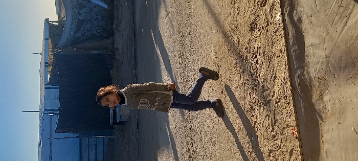 A young girl, approximately 6-10 years old, with curly brown hair, walks from right to left across an uneven dirt and sand path. She wears a light brown knit sweater with "HENA" or "HYNA" and two interconnected shapes (possibly hearts or glasses) printed in white on the front, a black turtleneck beneath, dark blue jeans, and dark brown sandals. Her gaze is directed towards the left. Long shadows are cast on the ground from the right, indicating low-angle sunlight. In the background, several temporary shelters are visible, consistent with an encampment in Khan Yunis, Palestinian Territory. These structures are made from white tarpaulins, some with blue stripes, stretched over frames. One shelter features a dark-colored tarpaulin with white patterns. A grey bucket or basin rests on the ground near the background structures. The sky is clear and light blue.