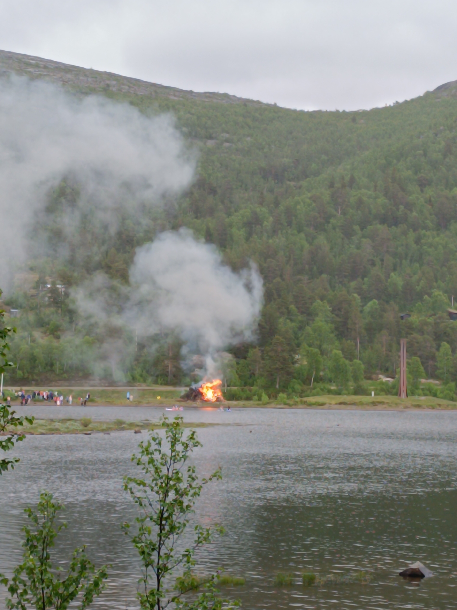 The image depicts a wide shot of a body of water with a fire and smoke billowing from a pyre on the shore. A group of people are gathered on the shore near the fire. The background features a densely wooded hillside under a cloudy sky. The location is identified as Hovden, Norway. The fire appears to be large and actively burning, with significant smoke production. The people on the shore are indistinct and appear to be observing the event. The water in the foreground is rippled, suggesting a gentle breeze. The overall scene suggests an outdoor event or celebration.