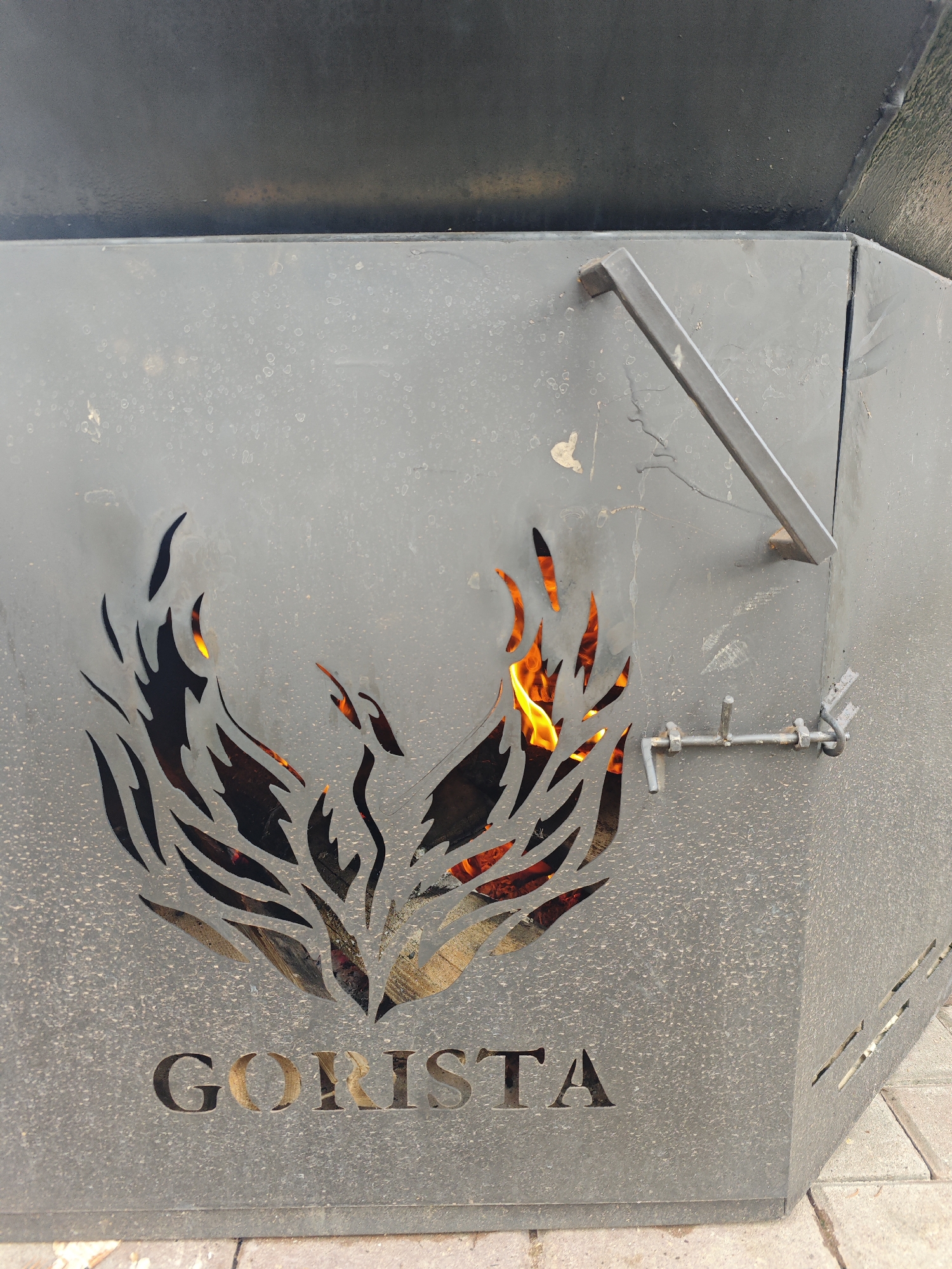 A medium shot captures a close-up of a dark grey, metal fire pit with a decorative cutout of a phoenix or bird of prey in flames. The fire pit has the word "GORISTA" cut out in large letters at its base. A metal latch is visible on the right side of the fire pit, securing a hinged door. The background is blurred, suggesting an outdoor setting with some paved ground visible at the bottom. The overall scene appears to be located in an outdoor urban environment, possibly a park or recreational area.
