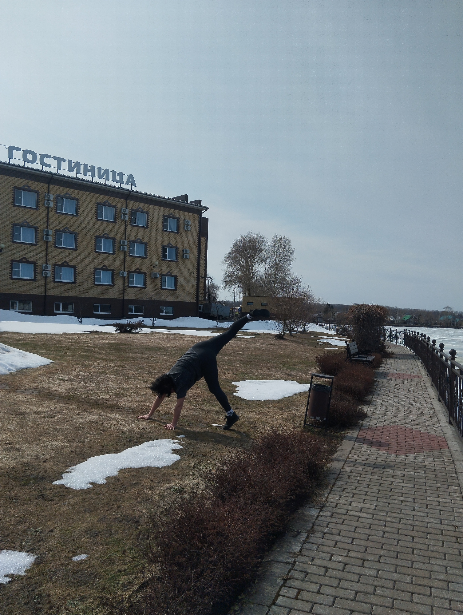 A person is performing a yoga pose in a grassy area with patches of snow, with the "Gostinitsa" (Hotel) building visible in the background, situated along a paved pathway beside a body of water in Votkinsk, Russia. The person is wearing dark clothing and is upside down with one leg extended upwards. A metal trash can is next to a hedge along the pathway. The sky is overcast.