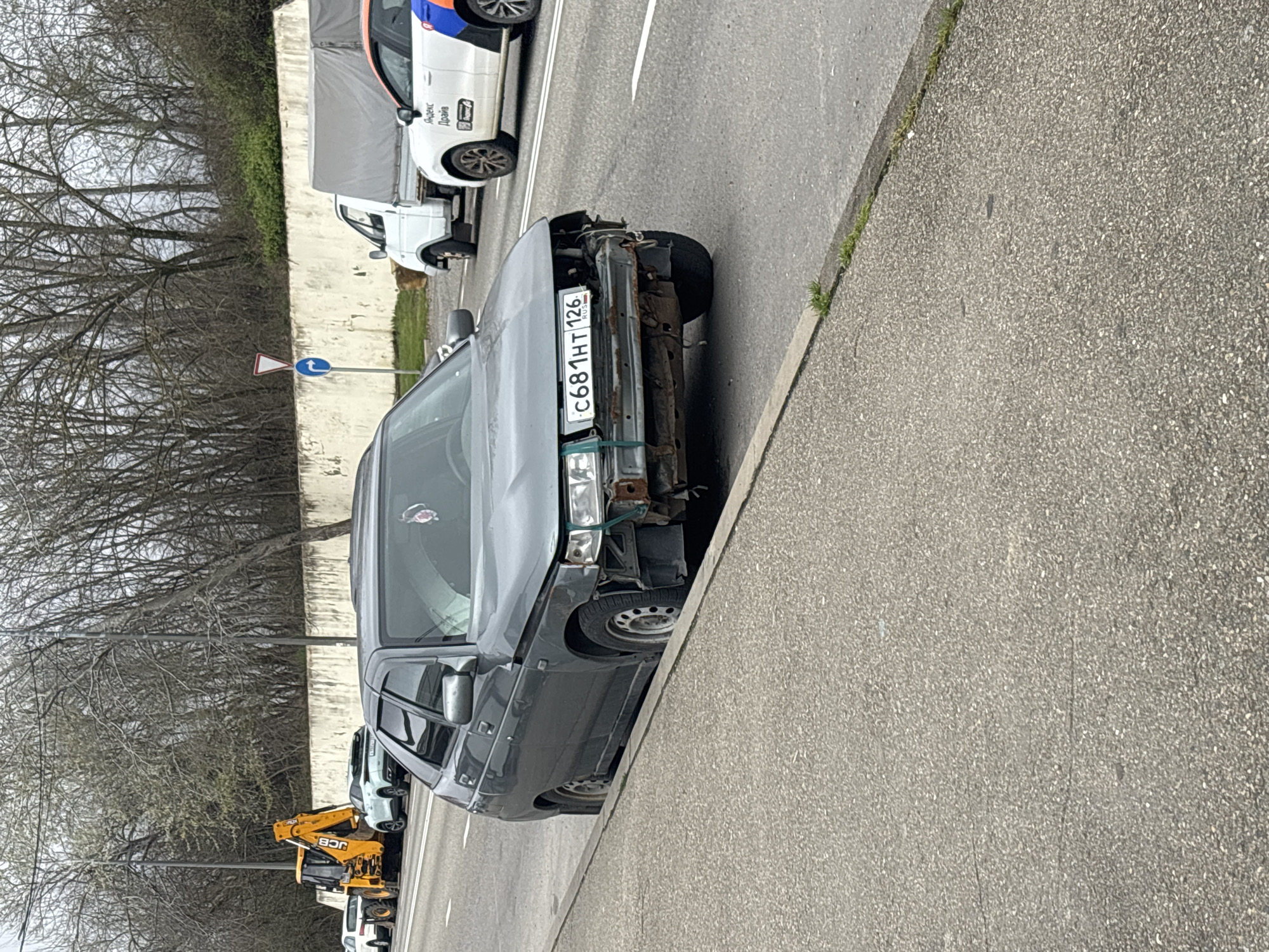 The image depicts a damaged gray Lada car parked on the side of a road in Sochi, Russia. The front of the car is heavily damaged, with a missing bumper and exposed internal components. The car's license plate reads "C681HT 126 RUS." Other vehicles, including a white van and a yellow JCB excavator, are parked further down the road. Trees and buildings are visible in the background. The sidewalk is visible adjacent to the damaged car, and the road appears to be a paved surface.