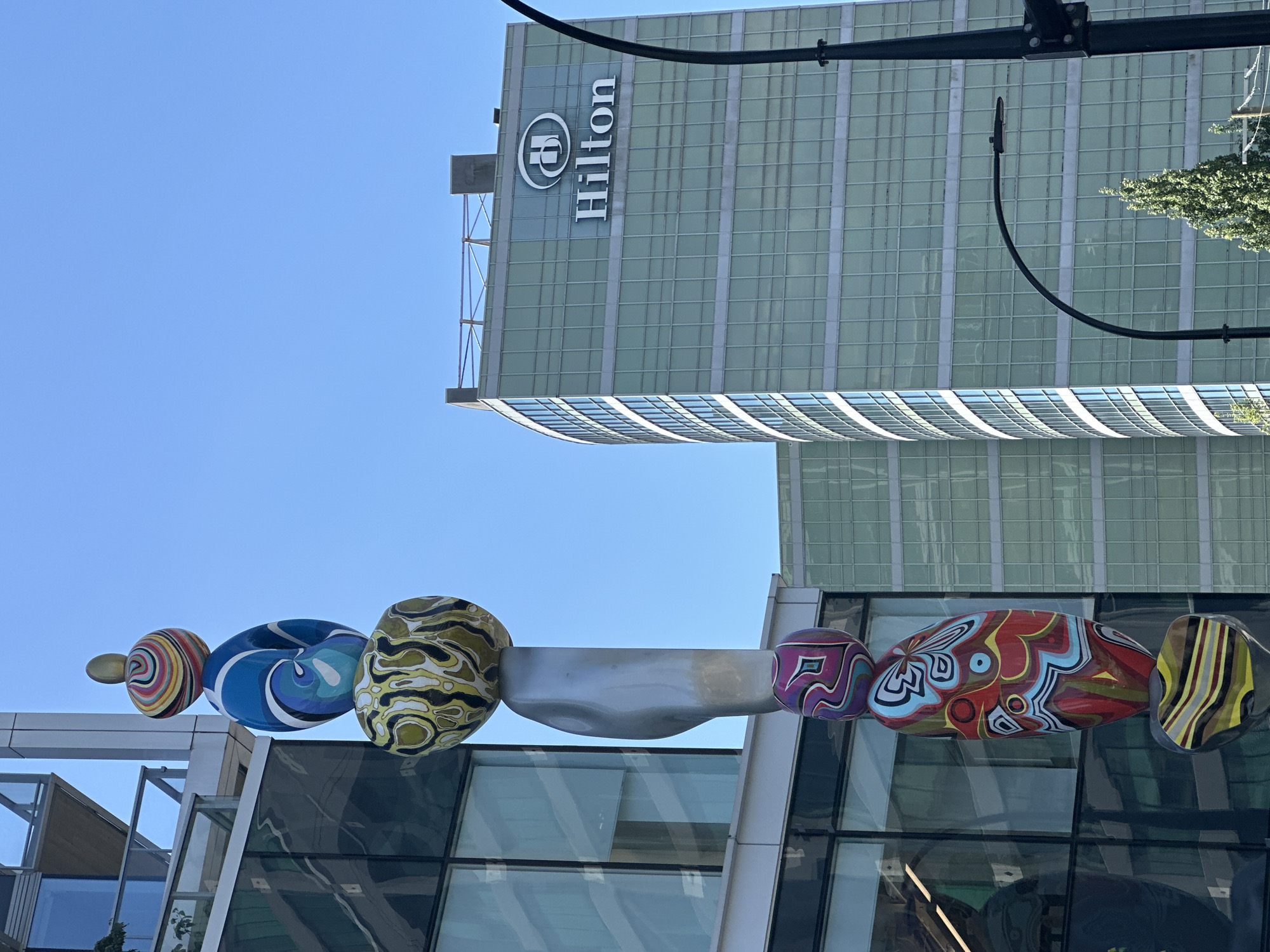 A public art sculpture, comprised of several brightly colored, bulbous shapes stacked vertically on a metallic base, stands in front of a modern building with a glass facade. The sculpture is predominantly in the left half of the frame, with the building occupying the right and upper portions. To the right of the sculpture and further back, a taller, curved building is visible, featuring a large "Hilton" logo in white lettering on its upper section. The sky is clear and blue, indicating daylight. The scene appears to be an urban environment. The sculpture's style and the architecture suggest it is located in a city. The Hilton hotel is a prominent landmark.