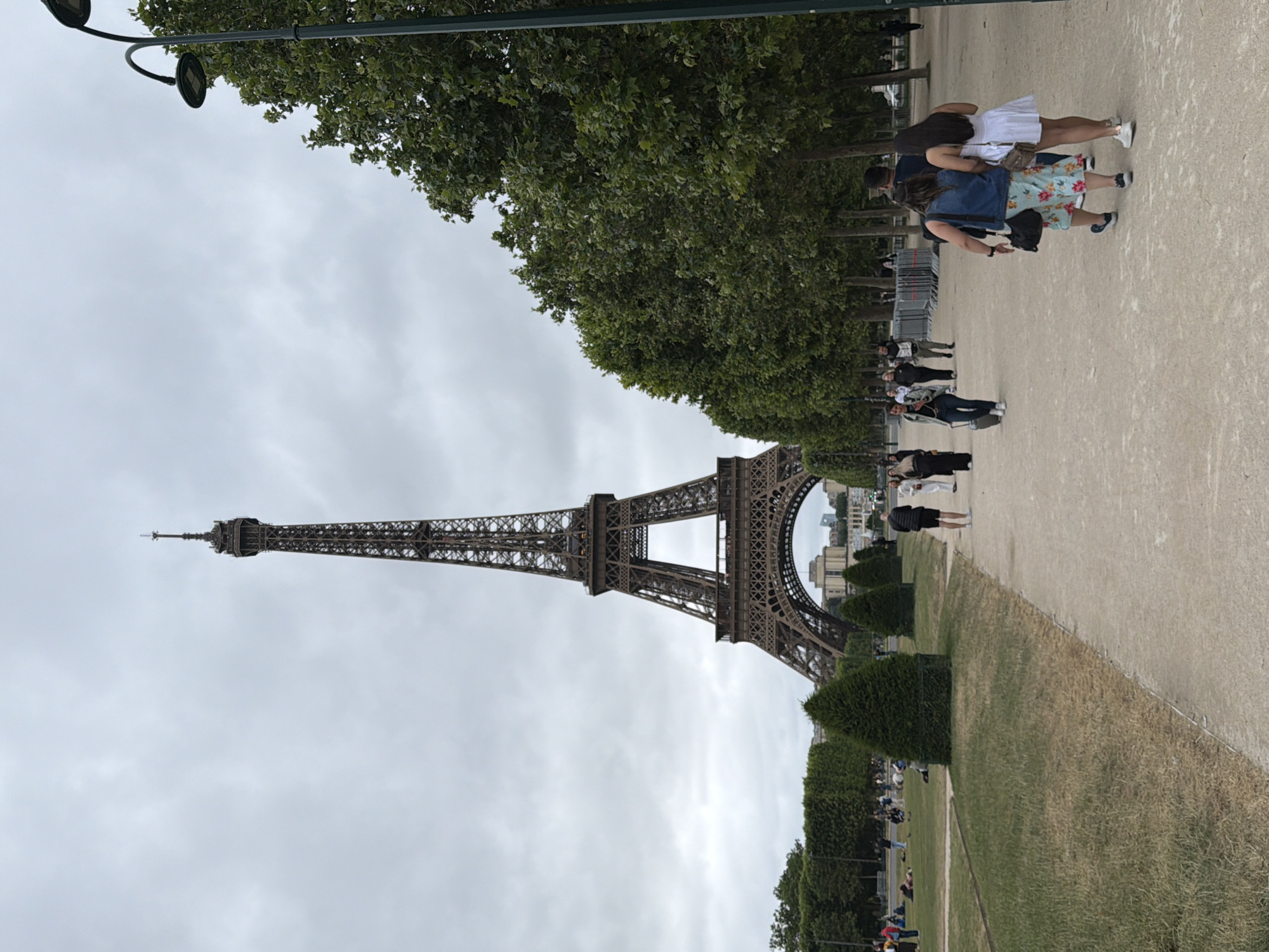 This image captures a daytime scene at the base of the iconic Eiffel Tower in Paris, France, under an overcast sky. The majestic iron lattice structure of the Eiffel Tower dominates the left side of the frame, rising vertically into the cloudy sky.
The scene is set within a public park, likely the Champ de Mars, characterized by broad walking paths and expansive green spaces. A wide, light-colored gravel or paved pathway stretches from the foreground towards the distance on the right. Along the left edge of this path, a dense line of lush green trees forms a natural canopy, obscuring the sky directly above the path in some areas. On the right side of the path, a manicured grassy lawn with neatly trimmed hedges separates it from another, larger grassy area where more people are visible.
Numerous people are scattered throughout the park, indicating a popular tourist destination. In the mid-ground on the path, a group of individuals is seen standing; one person wears a white dress, and another sports a floral skirt and a denim jacket. Further down the path and on the grass, other visitors are walking, standing, or sitting, observing the landmark and enjoying the surroundings. Temporary barriers are visible near some of the groups of people.
In the upper left corner, parts of a street lamp structure are visible, extending over the tree line. In the background, beyond the lower structure of the Eiffel Tower, glimpses of Parisian buildings can be seen under the same grey, cloudy sky. The overall atmosphere appears leisurely, with visitors engaged in sightseeing and enjoying the urban park setting. No visible text is clearly legible.
