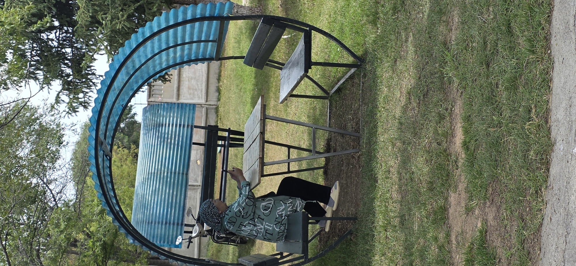 A woman wearing a headscarf and a green patterned jacket sits at a picnic table in an outdoor seating area. She is looking at her phone. The seating area is covered by a semicircular blue corrugated roof supported by black metal framing. A black bench seat is visible on the left side of the table and another on the right side. A motorcycle is parked behind the seating area. The ground is covered in grass and dirt. In the background, a concrete fence and trees are visible.