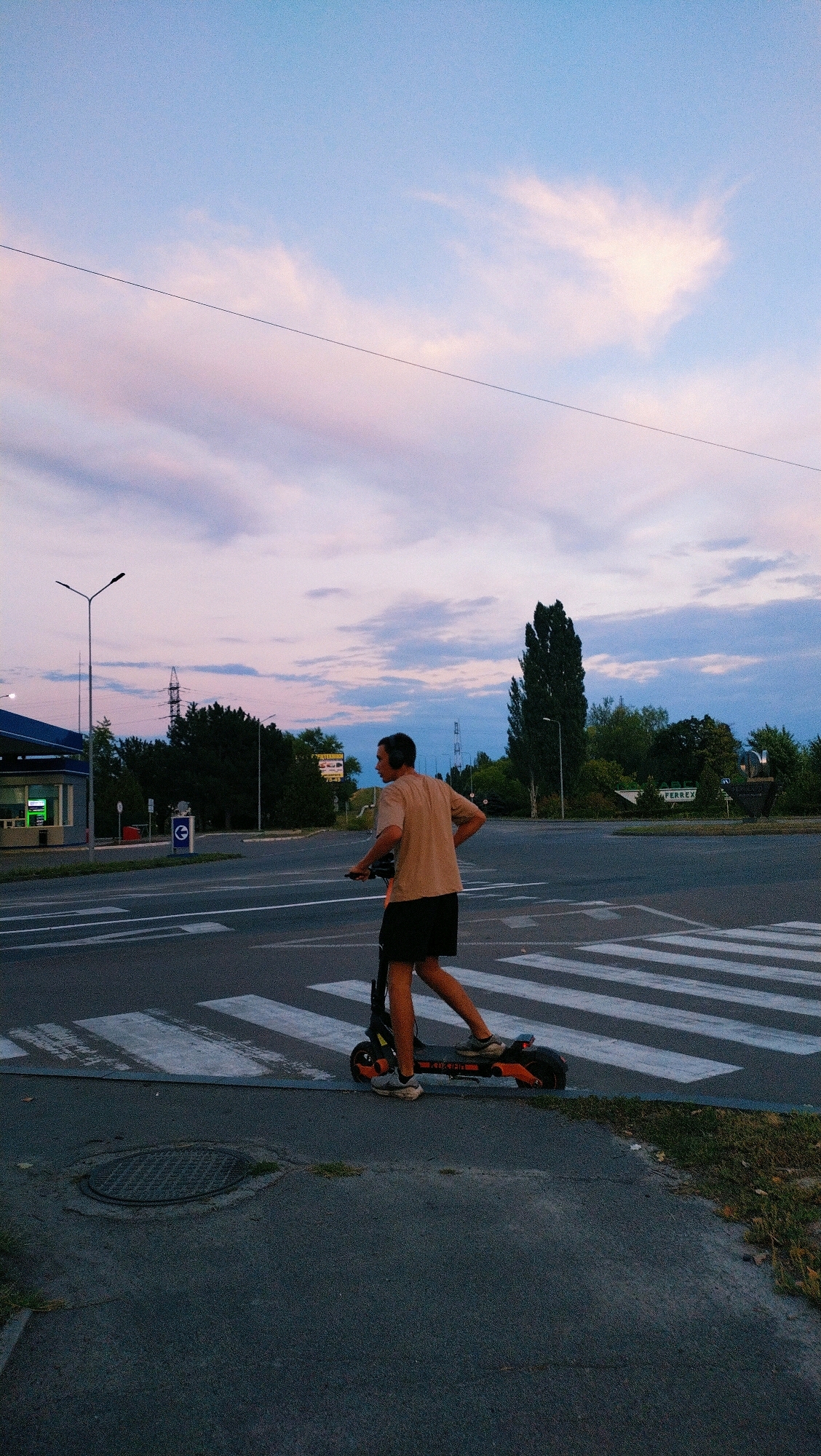A young man wearing headphones, a tan t-shirt, and black shorts rides an orange and black electric scooter across a crosswalk. The scooter has the word "OXELO" visible on its deck. The man is looking to his left, away from the camera. The sky is a twilight blue with streaks of pink and white clouds. In the background, there are trees, streetlights, power lines, a gas station, and buildings. The image appears to be taken in the city of Horishni Plavni, Ukraine.