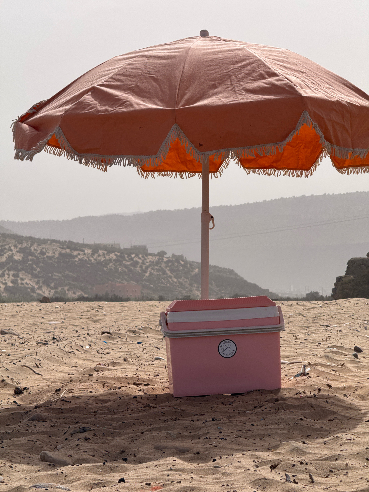 A pink cooler box sits on a sandy beach in Tamri, Morocco, under a large, peach-colored beach umbrella with white fringe. The umbrella is open and casts a shadow over the cooler. The beach is littered with small pieces of debris. In the background, rolling hills with sparse vegetation and some buildings are visible under a hazy sky. The overall impression is a serene beach scene.
