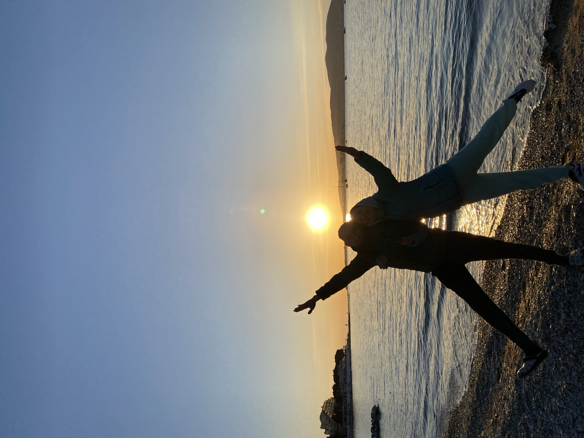 The image captures a vibrant coastal scene at either sunrise or sunset, featuring two individuals striking a playful pose on a pebbly shoreline. The photograph appears to be rotated 90 degrees clockwise.
The main subjects are two people, silhouetted against the brilliant, low-hanging sun. They are positioned centrally in the frame, both extending their arms horizontally and lifting one leg, creating a dynamic and balanced stance, as if mimicking flight or dancing. The person on the left wears dark clothing, while the one on the right is dressed in lighter, possibly teal-colored, athletic wear. Their cheerful postures suggest a moment of joy and carefree fun.
In the background, a vast body of water, likely a sea or bay, extends to the horizon, its surface shimmering with reflections of the golden sun. The immediate foreground features a textured, pebbly or rocky beach where the subjects are standing. Across the water, a distant landmass or low mountain range is visible, silhouetted against the bright sky. Faint architectural details, possibly buildings of a coastal town, can be discerned along the distant shore, and a few small boats dot the water.
The time of day is clearly either sunrise or sunset, characterized by the sun's low position and the dramatic backlighting. The sky transitions from a clear, deep blue at the top to warm golden and orange hues around the sun. The weather appears clear and calm, with gentle waves on the water. The location, as per context, is Gelendzhik, Russia. There is no visible text in the image.