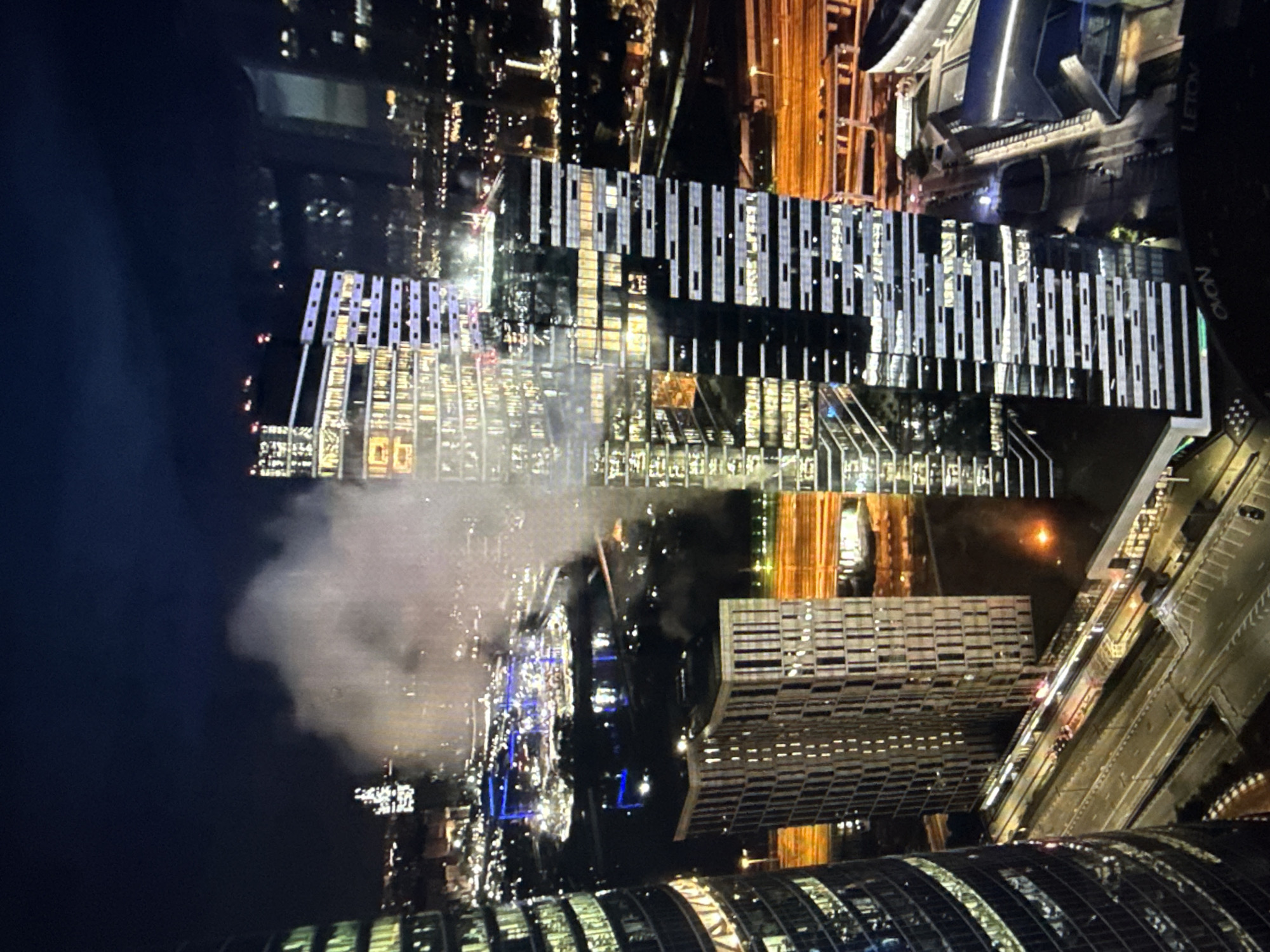 This is a nighttime, high-angle shot of a dense urban landscape, dominated by modern skyscrapers. A significant amount of white smoke or mist is obscuring a portion of the city in the middle ground, creating a diffused haze over a section of buildings and infrastructure. In the foreground and mid-ground, several tall, illuminated buildings are visible. One building on the left has a curved, dark facade with horizontal bands of light. Adjacent to it is a lighter-colored building with a grid-like facade, appearing to be under construction or having a unique architectural design. Another prominent skyscraper in the mid-right has a sleek, modern design with alternating dark and light horizontal bands, reflecting light.
Below the buildings, elevated roadways and street-level infrastructure are visible, illuminated by streetlights and vehicle headlights. The distant cityscape shows a dense collection of lights, suggesting a sprawling metropolitan area. The sky above is dark blue, indicative of nighttime. The overall impression is one of a dynamic, illuminated city, with the unusual element of the prominent smoke plume adding a dramatic and mysterious quality to the scene. The location is identified as Unknown, Unknown.