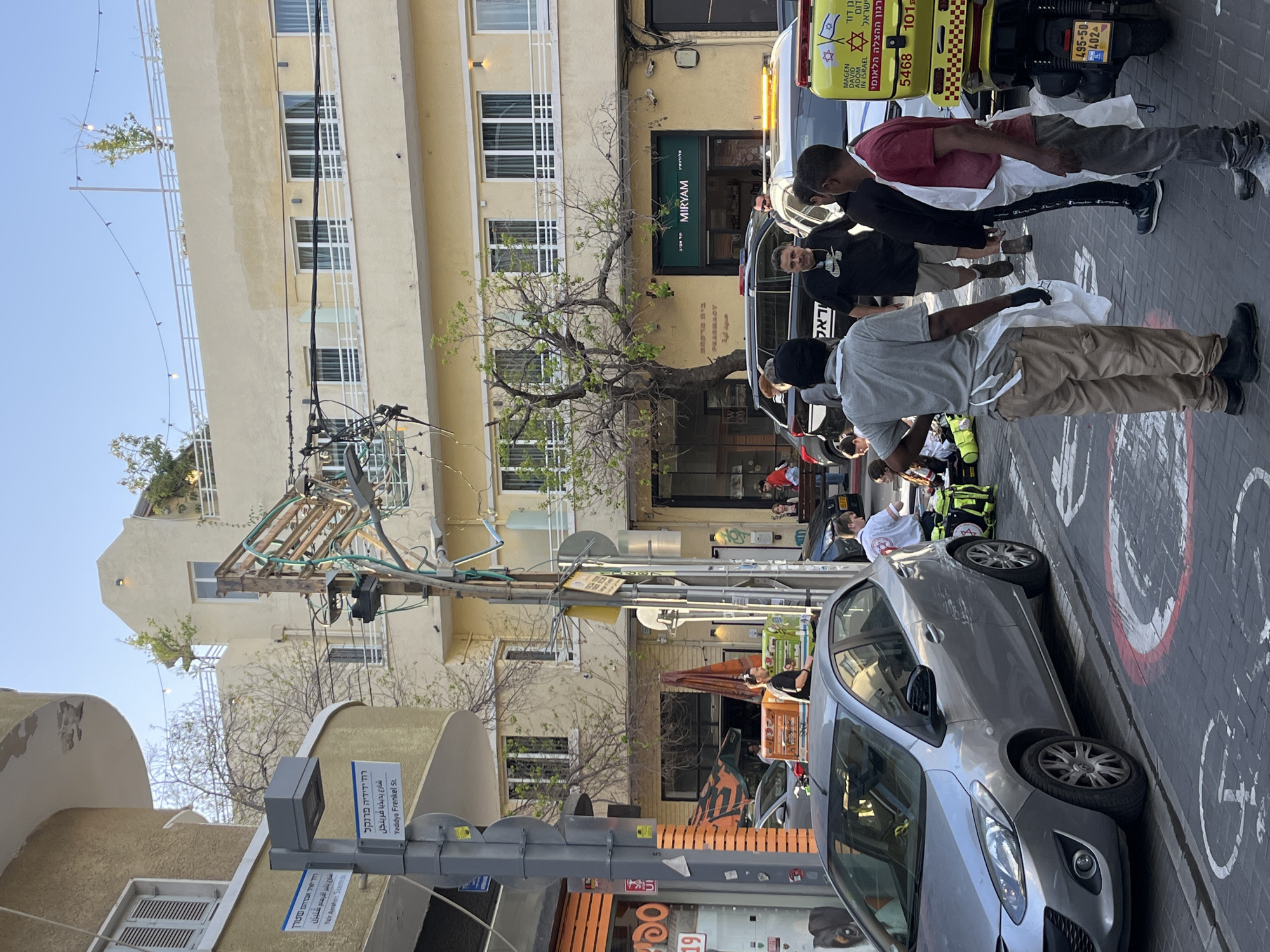 The image depicts a street scene in Tel Aviv, Israel. A Magen David Adom ambulance is visible on the right. Several people are standing near the ambulance, some wearing white aprons. A grey car is parked on the side of the street, partially obscuring a sign that reads "Yedidya Frenkel St." in Hebrew and English. The street is paved with brick. Buildings line the street, with trees scattered throughout. A sign for a pharmacy named "Miryam" is visible. The street is lined with retail shops.