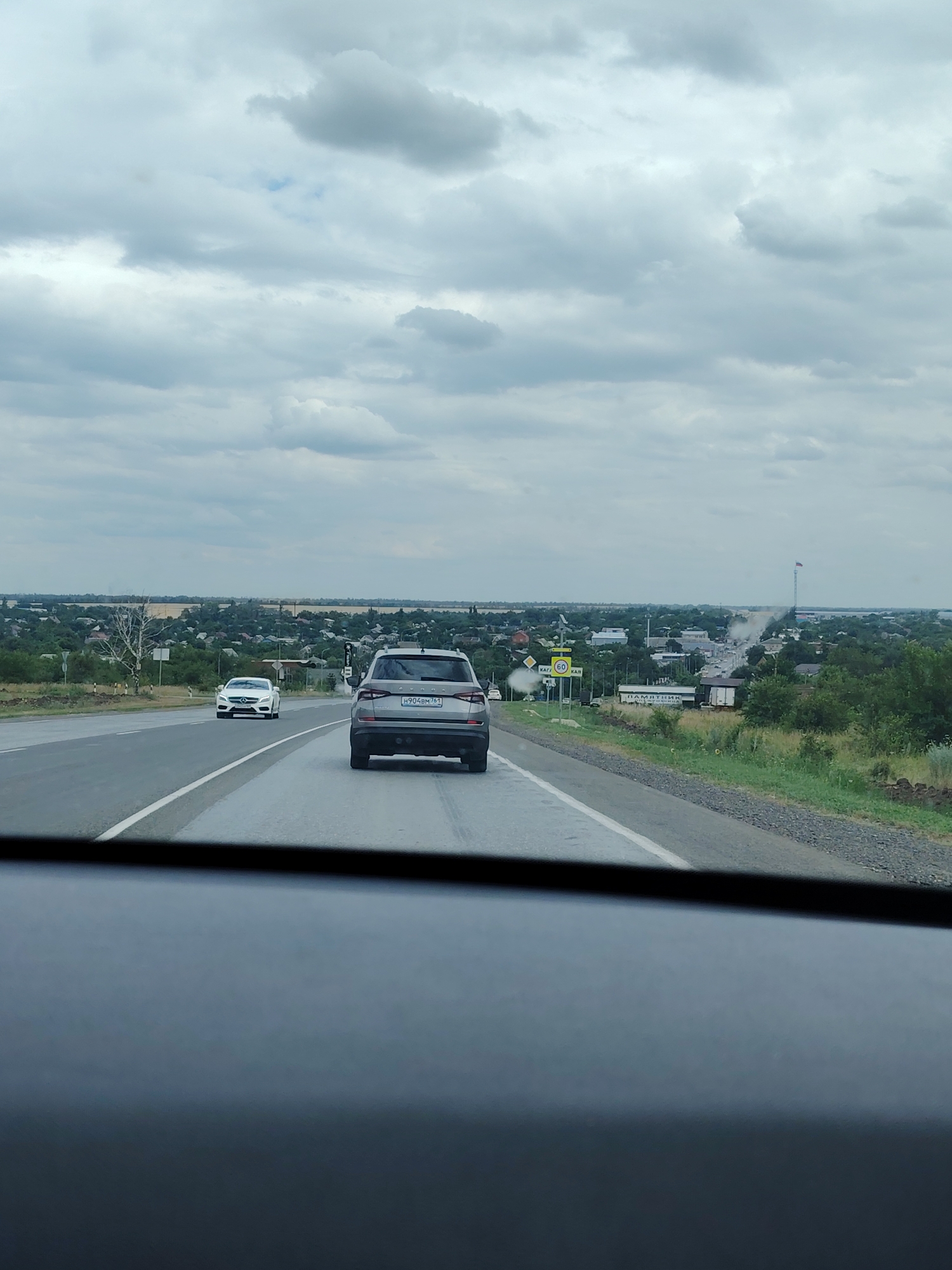 The image depicts a view from inside a vehicle traveling on a highway towards the city of Kagal'nitskaya, Russia. In the foreground, the dashboard of the vehicle is visible. On the highway, two cars are traveling in the same direction. A dark grey SUV is in the right lane, and a white sedan is in the left lane. In the distance, Kagal'nitskaya is spread out with numerous buildings and trees. A tall flagpole with a Russian flag is prominently visible in the city. The sky is overcast with heavy clouds. Road signs, including a speed limit sign for 60, are also present along the side of the highway. The overall scene suggests a typical daytime drive on a highway approaching a town.