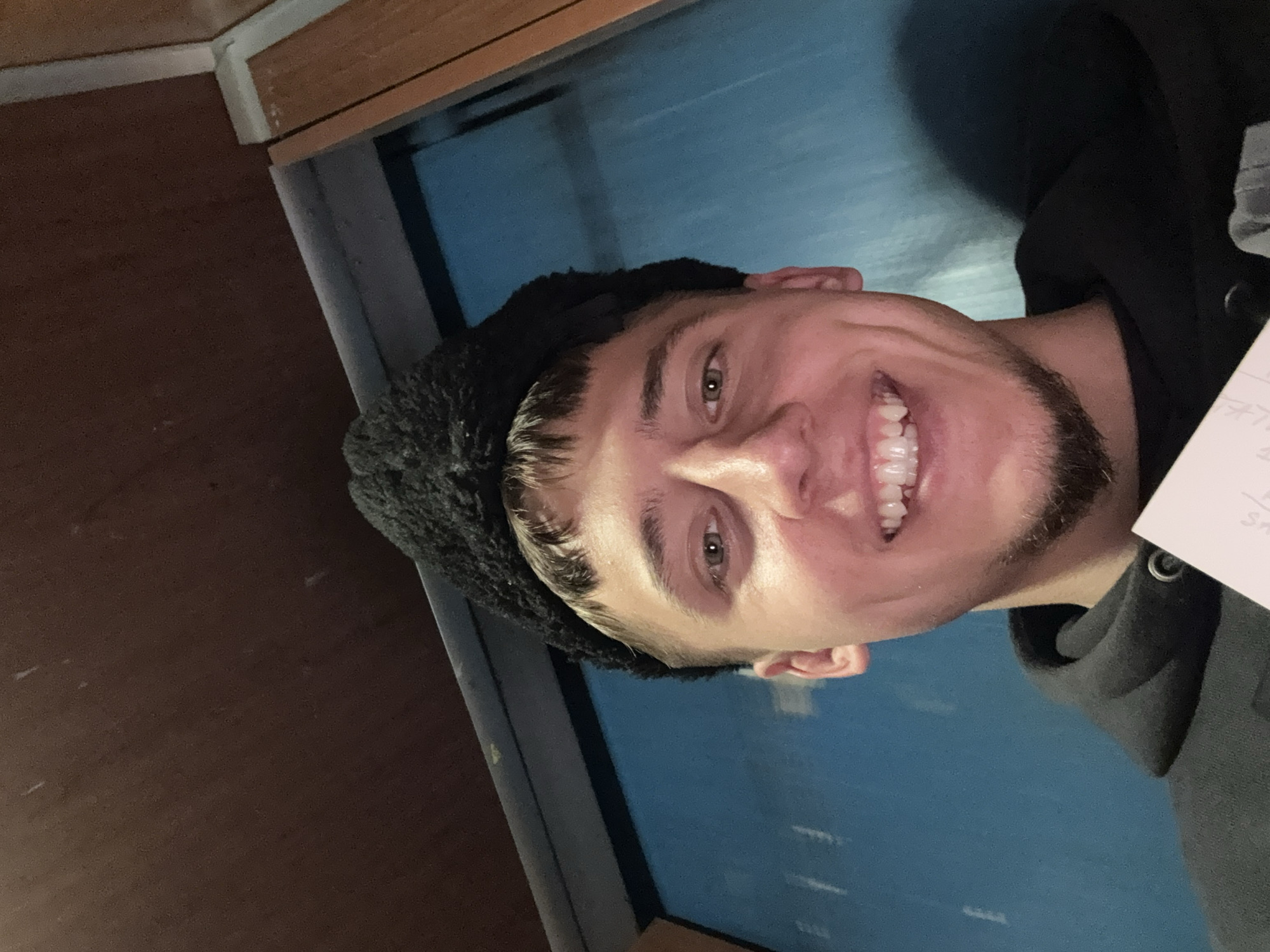 A close-up photograph shows a smiling man with light skin wearing a black fuzzy hat and a gray hooded sweatshirt. He has short, dark brown hair and a beard. He holds a piece of paper in his right hand. The background features a blue wall or window, and a portion of a brown wooden surface is visible in the upper left corner. The image appears to be taken indoors. The location in Sofia, Bulgaria is unknown.