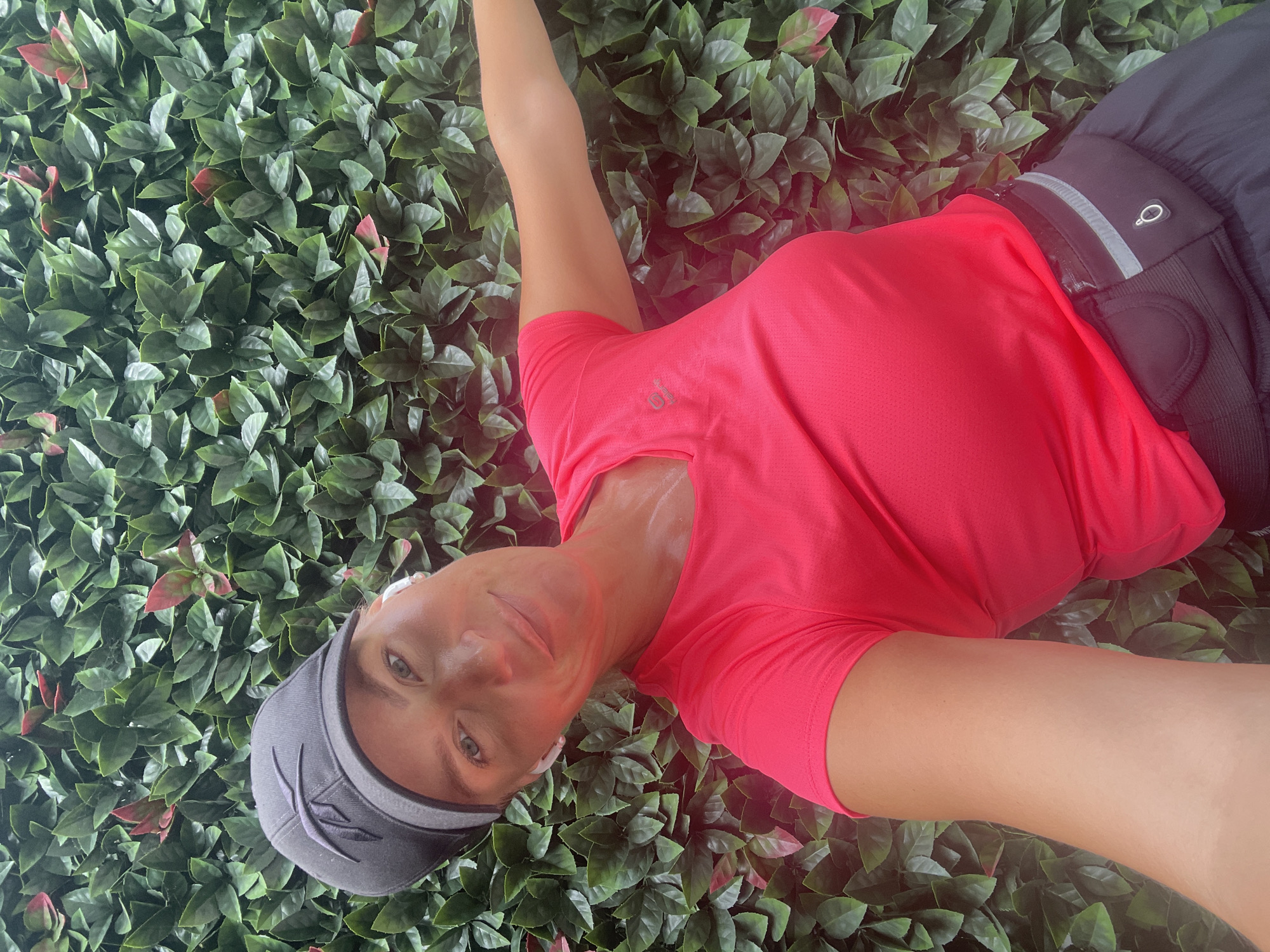 A woman wearing a pink t-shirt, a black baseball cap, and black shorts is photographed outdoors against a backdrop of green artificial foliage. She is also wearing white wireless earbuds and a black fanny pack around her waist. The woman is smiling and looking towards the camera, with her right arm extended outwards. The image appears to be a selfie. The location is not specified in the image or its context.