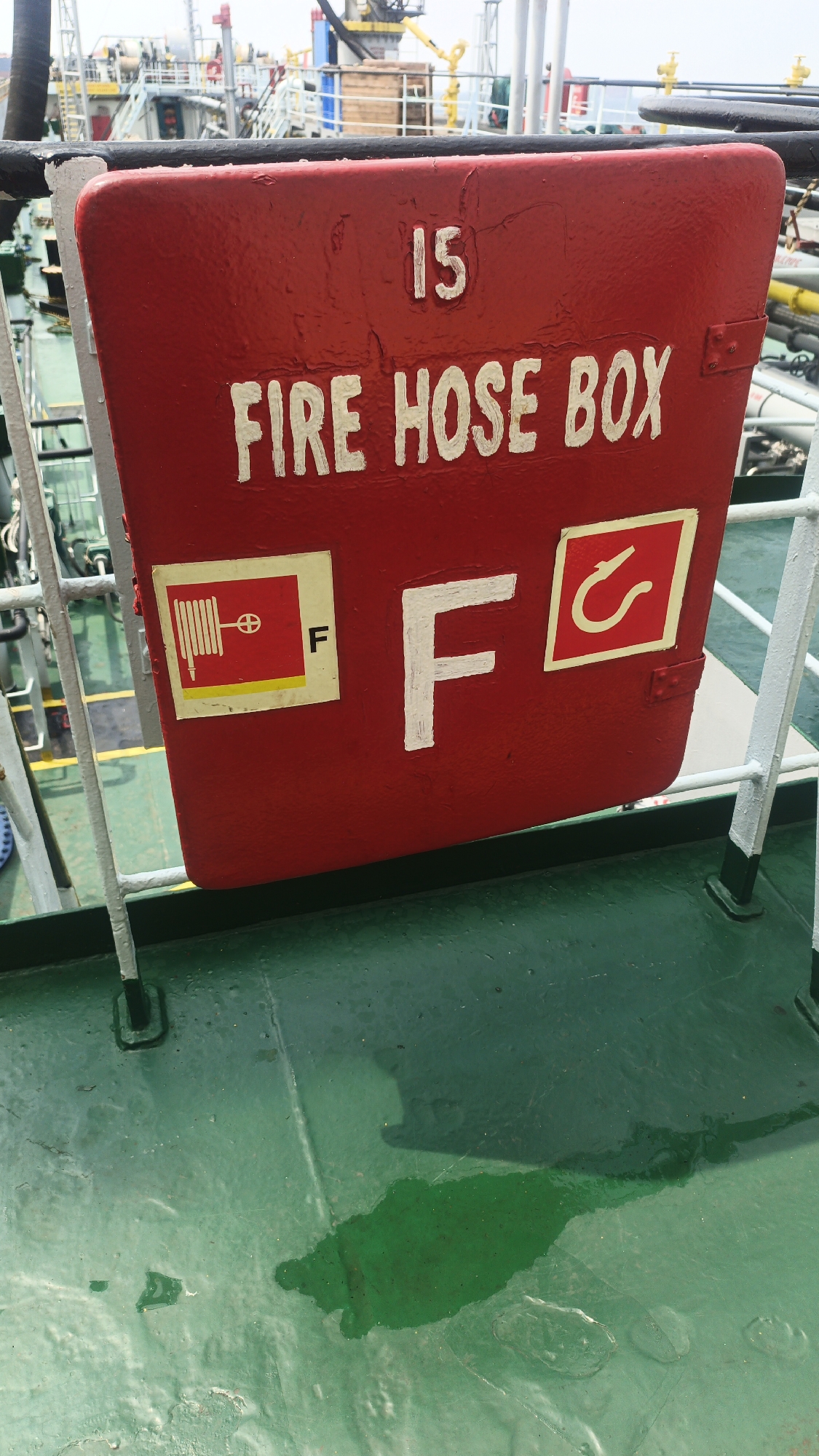 A red metal "FIRE HOSE BOX" with the number "15" painted in white on it is mounted on a railing above a green metal deck. The box has two square symbols on it, one on the left with a drawing of a hose and nozzle and the letter "F", and another on the right with a hook symbol. A large "F" is painted in white in the center of the box. The green deck shows signs of wear and a large spill of green liquid. In the background, other ships and industrial structures are visible, suggesting a port or industrial area. The image's perspective and the presence of ships suggest it might be located in a maritime setting, possibly a port in Singapore.