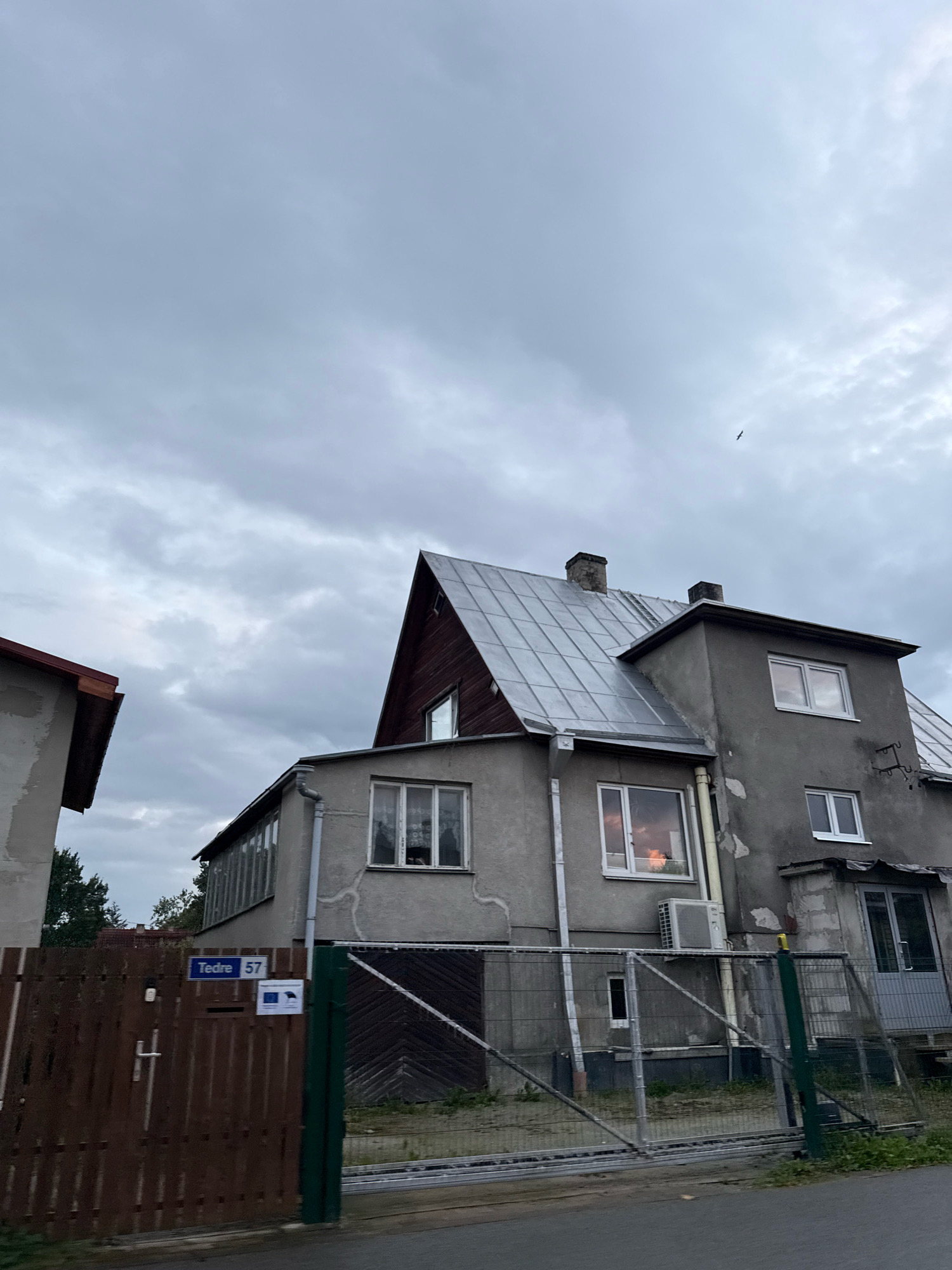 This is a vertical, eye-level, medium shot of a residential building on a cloudy day in Tallinn, Estonia. The building is a multi-story structure with a grey, textured exterior and a grey metal roof. The building features several windows, some of which reflect the sky and warm interior lights. An air conditioning unit is visible mounted on the exterior wall. A wooden gate with a metal latch is in the foreground, to the left, with a blue street sign reading "Tedre 57" and two small square signs attached to it. A metal fence runs in front of the building. In the sky, a bird is silhouetted against the clouds.