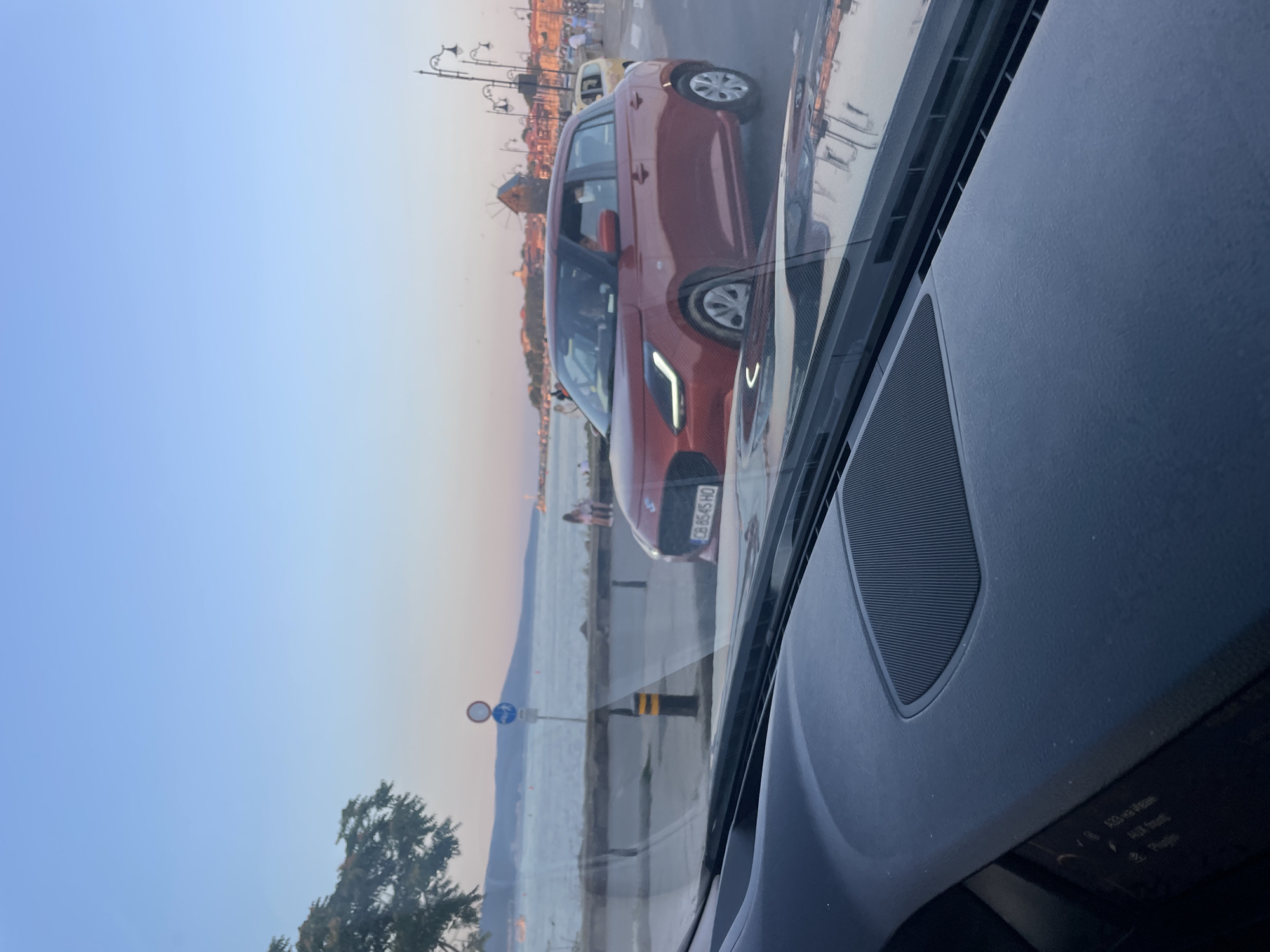 The image captures a view from inside a car, looking through the windshield at an outdoor scene in Nesebar, Bulgaria. The foreground features the car's dashboard, with a speaker grille visible on the right side. A portion of the infotainment screen is also visible on the lower left, displaying text that includes "AUX Volume" and "Plugins".
Outside the car, a red hatchback is the most prominent subject, parked or slowly moving on a street parallel to a waterfront. The car has a license plate that reads "CB 8545 HO". Beyond the street, a body of water, likely the Black Sea, stretches towards the horizon, with distant hills and buildings visible. The sky is a clear, pale blue, transitioning to a lighter hue near the horizon, suggesting it is either early morning or late afternoon.
Along the waterfront, a few people are visible, some walking and others seated. Streetlights with multiple lamps line the street, and a traffic sign with a red circle and a blue sign below it is mounted on a pole. In the middle ground, a tree with sparse foliage is on the left, and a quaint, old-fashioned windmill is visible in the background on the right, a characteristic feature of Nesebar. The overall impression is a serene, coastal urban environment.