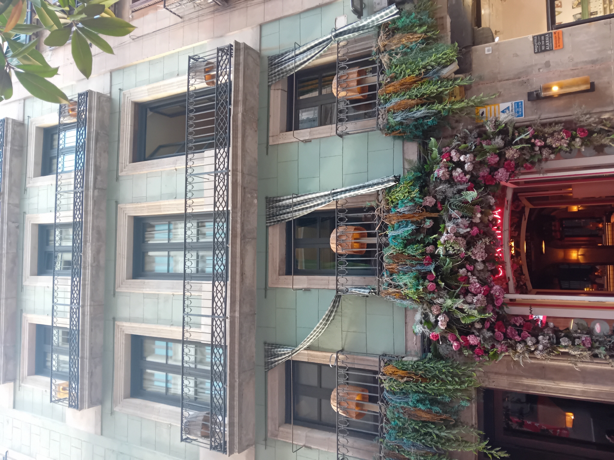 The image shows the exterior of a building in Barcelona, Spain. The building has a light blue facade and several windows with wrought iron balconies. The windows are adorned with striped awnings and hanging plants. The entrance to a restaurant or bar is decorated with flowers and a neon sign. Security information is visible on a nearby wall. A tree branch appears in the top left corner of the image.