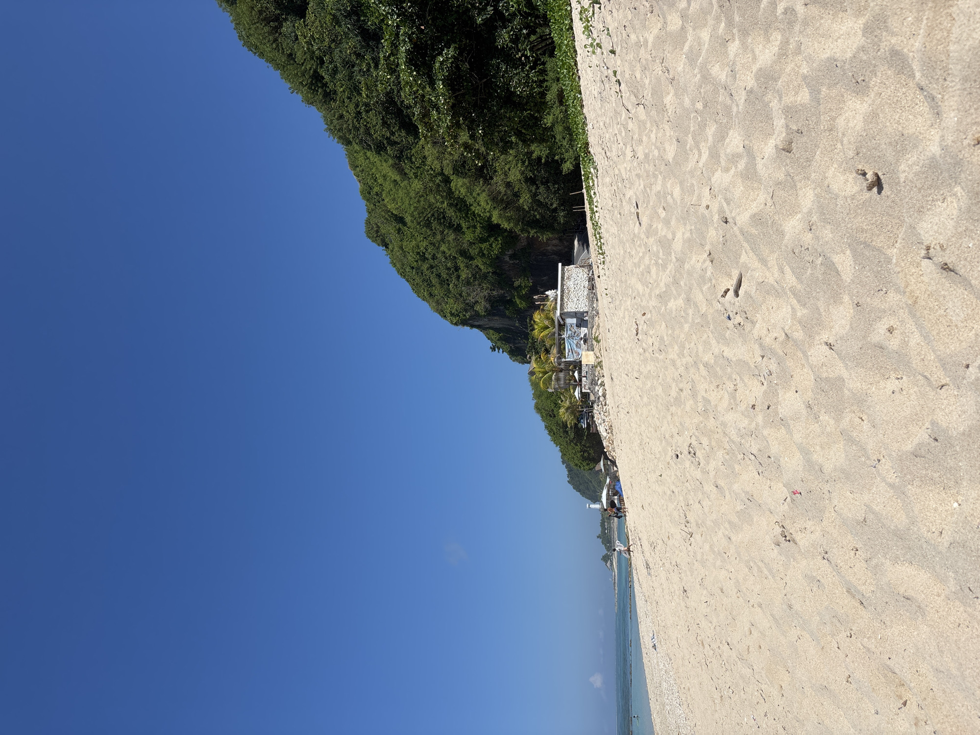 The image captures a beach scene in Kutuh, Indonesia, presenting a clear blue sky that occupies a substantial portion of the frame. To the right, a sandy beach slopes toward a strip of water along the bottom edge of the image.
Adjacent to the beach is a densely vegetated cliff face. Towards the middle ground, structures are nestled along the cliff's edge, suggesting a resort or private dwellings. Several people are visible along the shoreline. The sand texture indicates footprints and disturbances.