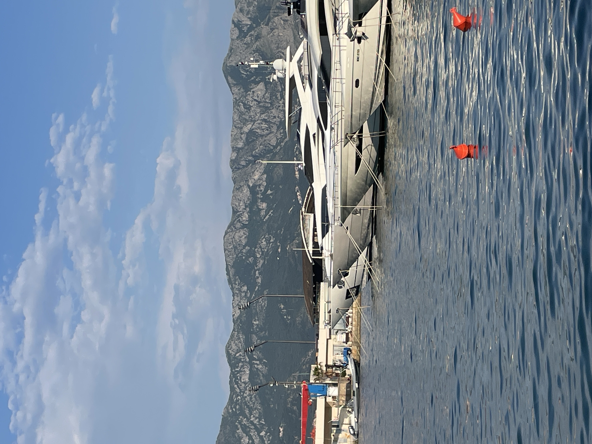 The image presents a view of a marina in Budva, Montenegro. A large white yacht is docked on the right side of the image. Two orange buoys float in the water near the yacht. A mountain range provides the backdrop, with a blue sky dotted with white clouds above. Closer to the dock, one can see streetlights and some harbor-related infrastructure, including a red and blue crane.