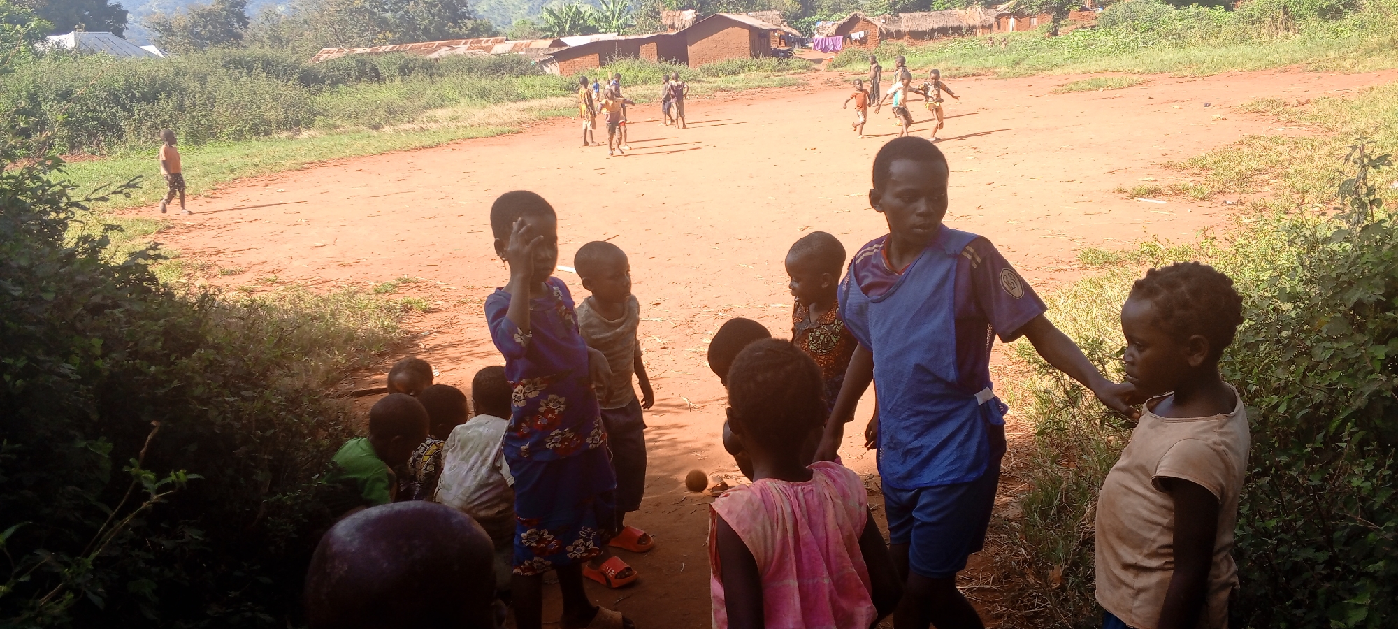 The image depicts a group of children in Ngalula, Democratic Republic of the Congo. In the foreground, several children are gathered near vegetation. One wears a blue floral-patterned dress, and another is in a blue shirt with shorts. One child wears a pink top.
In the background, a larger group of children are playing on a dirt field near several simple, mud-brick houses with corrugated iron roofs. Vegetation surrounds the field and the houses. A lone child walks from left to right in the background of the field. The sky is partly obscured by trees and hills in the distance. The light suggests it is daytime, possibly mid-morning or mid-afternoon.