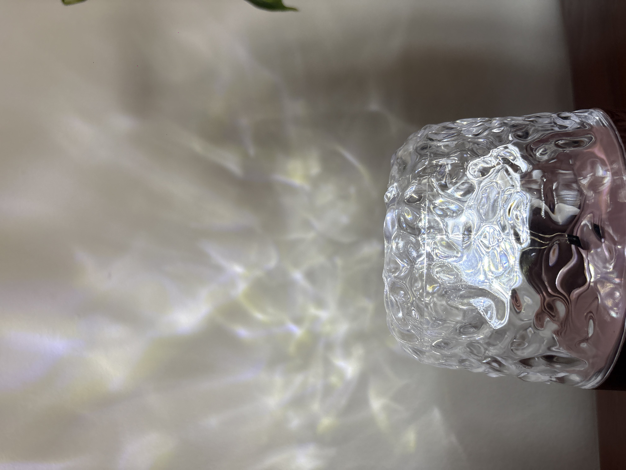 The image presents a close-up view of a clear, textured object, likely made of glass or plastic, situated on the right side of the frame. The object possesses an uneven, rippled, or bubbly surface that interacts with light.
The most striking feature of the image is the intricate pattern of light projected onto the light-colored, possibly off-white or beige, background to the left. These patterns are iridescent and wave-like, primarily appearing in shades of white, yellow, and faint blue or purple, indicating the refraction and dispersion of light. The intensity and spread of these patterns suggest a strong light source, possibly sunlight, illuminating the textured object.
In the upper left corner, a small portion of a green leaf is visible, suggesting the presence of a plant nearby. A dark, indistinct area is present on the far right edge of the frame, behind the clear object. The image is captured from a slightly elevated perspective, looking down at the object and its cast light. There is no discernible activity or visible text in the image.