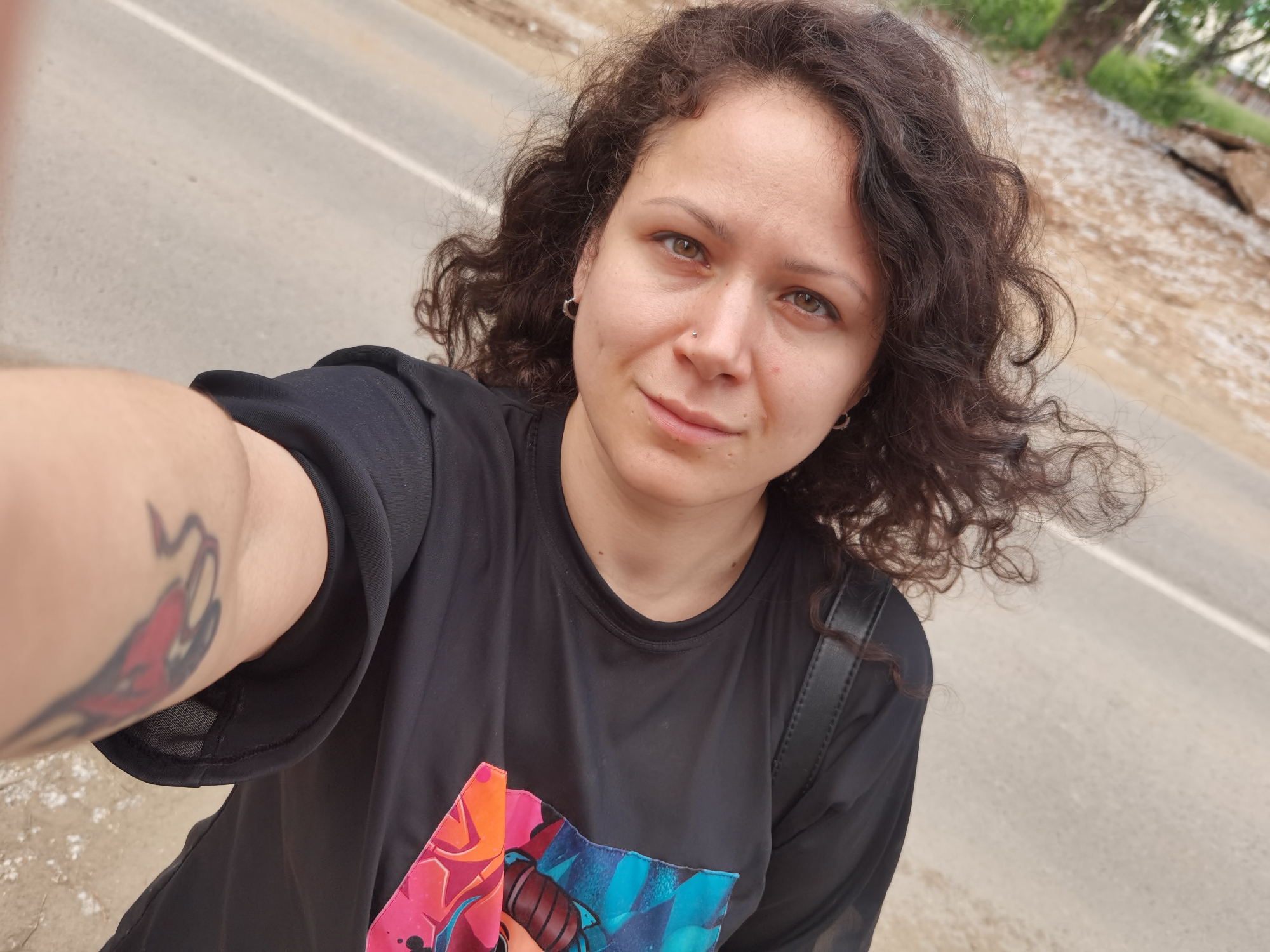 A woman with curly brown hair and light-colored eyes is taking a selfie outdoors. She is wearing a black t-shirt with a colorful graphic design on the front. She has a tattoo on her left arm and is wearing earrings and a nose ring. The background appears to be a street with a tree-lined area to the right. The image was taken in Chaikovsky, Russia.