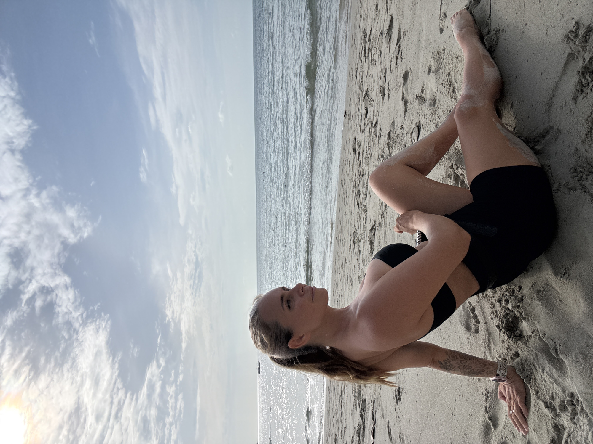 In Ko Samui District, Thailand, a woman with blonde hair in a ponytail sits on a sandy beach, facing to her left. She wears a black bikini top and black shorts. Her left arm rests on the sand, adorned with a tattoo and a watch. Her right hand is bent at the wrist and resting on her right leg. The ocean is visible behind her, with calm waves and a boat in the distance. The sky is partly cloudy, with the sun shining through the clouds in the upper left corner. The sand is textured with footprints.