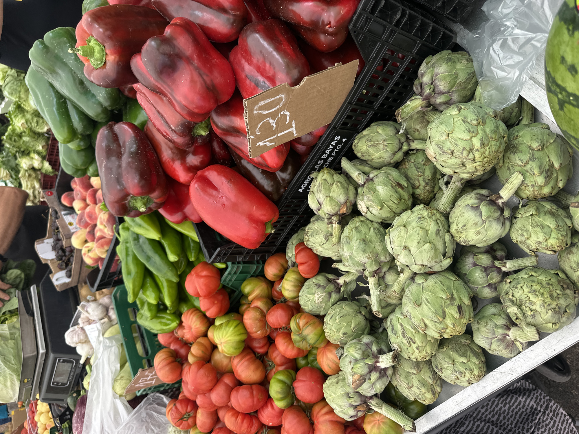 The image displays a vibrant outdoor market stall, showcasing a variety of fresh produce. In the foreground, large piles of red bell peppers and green bell peppers are prominently featured, with their bright colors contrasting against each other. To the right, a large quantity of green artichokes is stacked, revealing their distinct textured leaves and short stems. Below the bell peppers and artichokes, there is a substantial display of ripe and semi-ripe tomatoes, characterized by their ribbed, heirloom-like appearance, showing shades of red and green.
Further into the background and to the left, additional produce can be seen, including piles of green chili peppers, green leafy lettuce, and bunches of garlic. Peaches are visible behind some of the peppers. A watermelon is partially visible in the top right corner. The produce is arranged in black and green plastic crates, typical of market displays.
A piece of cardboard with "130" handwritten on it serves as a price tag among the red bell peppers. Another black crate has text printed on it: "AGRO LAS BAYAS" and "CHE PTO.4", suggesting a farm or distributor's name, possibly in a Spanish-speaking region. A digital weighing scale is visible on the left side, indicating commercial activity. A person's arm is partially visible on the far left. The scene is brightly lit, consistent with a daytime outdoor setting.