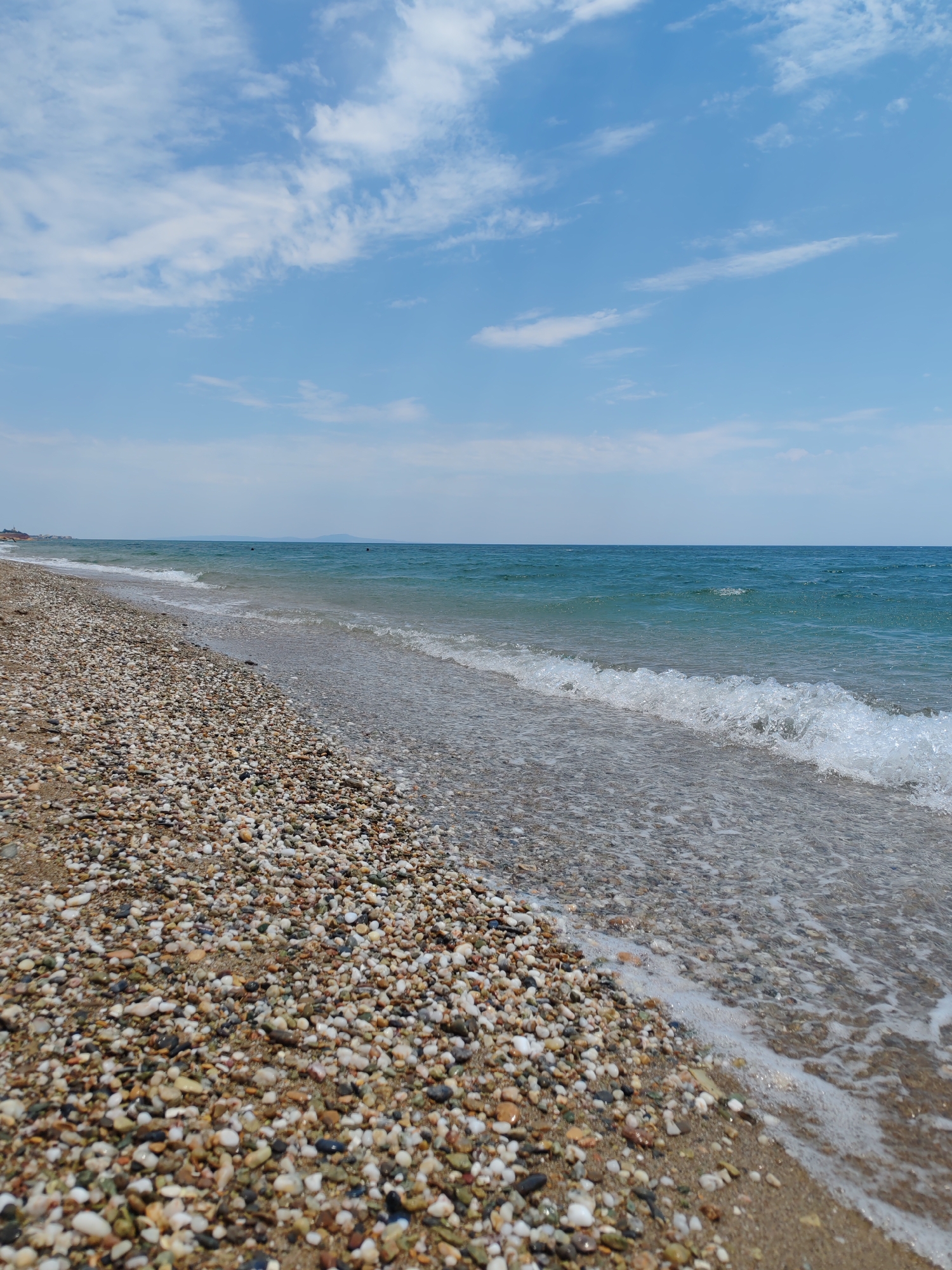 The image depicts a view of a pebble beach in Makri, Greece. The foreground is dominated by a dense scattering of small, multi-colored pebbles, interspersed with sand. A gentle wave is breaking on the shore, its white foam contrasting with the clear, turquoise water. In the middle ground, the sea stretches out to the horizon, appearing calm and expansive. The horizon line is relatively low, emphasizing the sky above, which is a vibrant blue with scattered white clouds. In the far distance, a faint, dark landmass or island is visible on the horizon. The overall scene is one of a tranquil coastal environment on a clear day. There are no people or specific man-made structures clearly visible in the immediate vicinity, suggesting a natural or sparsely populated beach area.