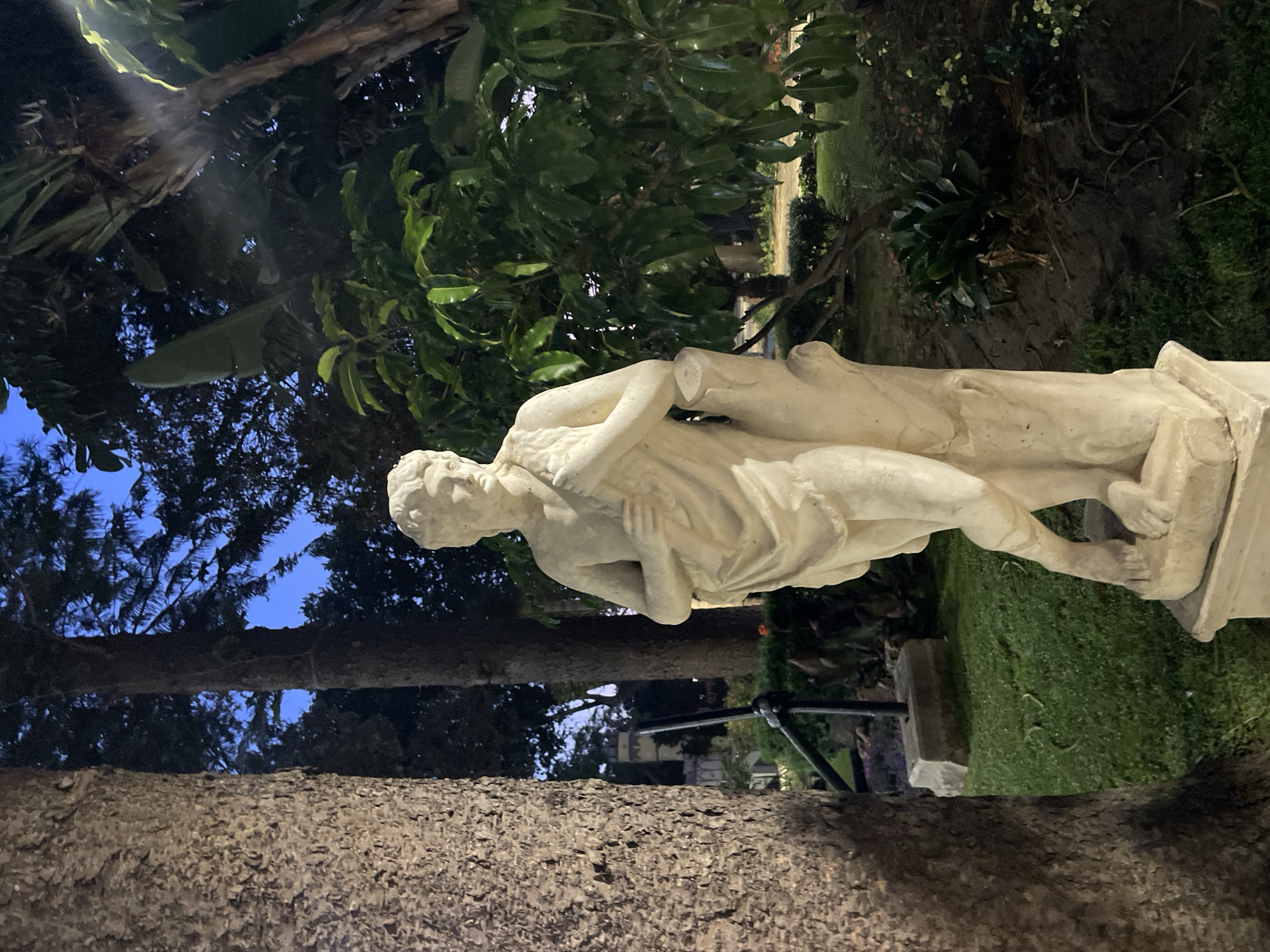A full shot of a classical-style statue of a male figure, carved from light-colored stone, stands outdoors at dusk in a garden. The statue is positioned slightly to the right of the frame, leaning against a tree trunk. The figure is draped in a cloth, with one arm bent and resting on the trunk. The statue is on a pedestal. In the background, large trees with dark foliage are visible, and the sky transitions from deep blue to lighter hues near the horizon, suggesting twilight. The ground is covered in grass, with some scattered plants and a dark, possibly dirt path, to the right of the statue. A building with a yellow turret and a distinctive architectural style is visible in the distance through a gap in the trees, situated to the left of the statue. The overall lighting suggests natural light diminishing, with some artificial lighting illuminating the grass in front of the statue, creating a soft glow. This scene is consistent with the surroundings of the gardens of the Hotel Mirador in La Línea de la Concepción, Spain, particularly the area facing the sea, where such statuary and lush vegetation are characteristic.