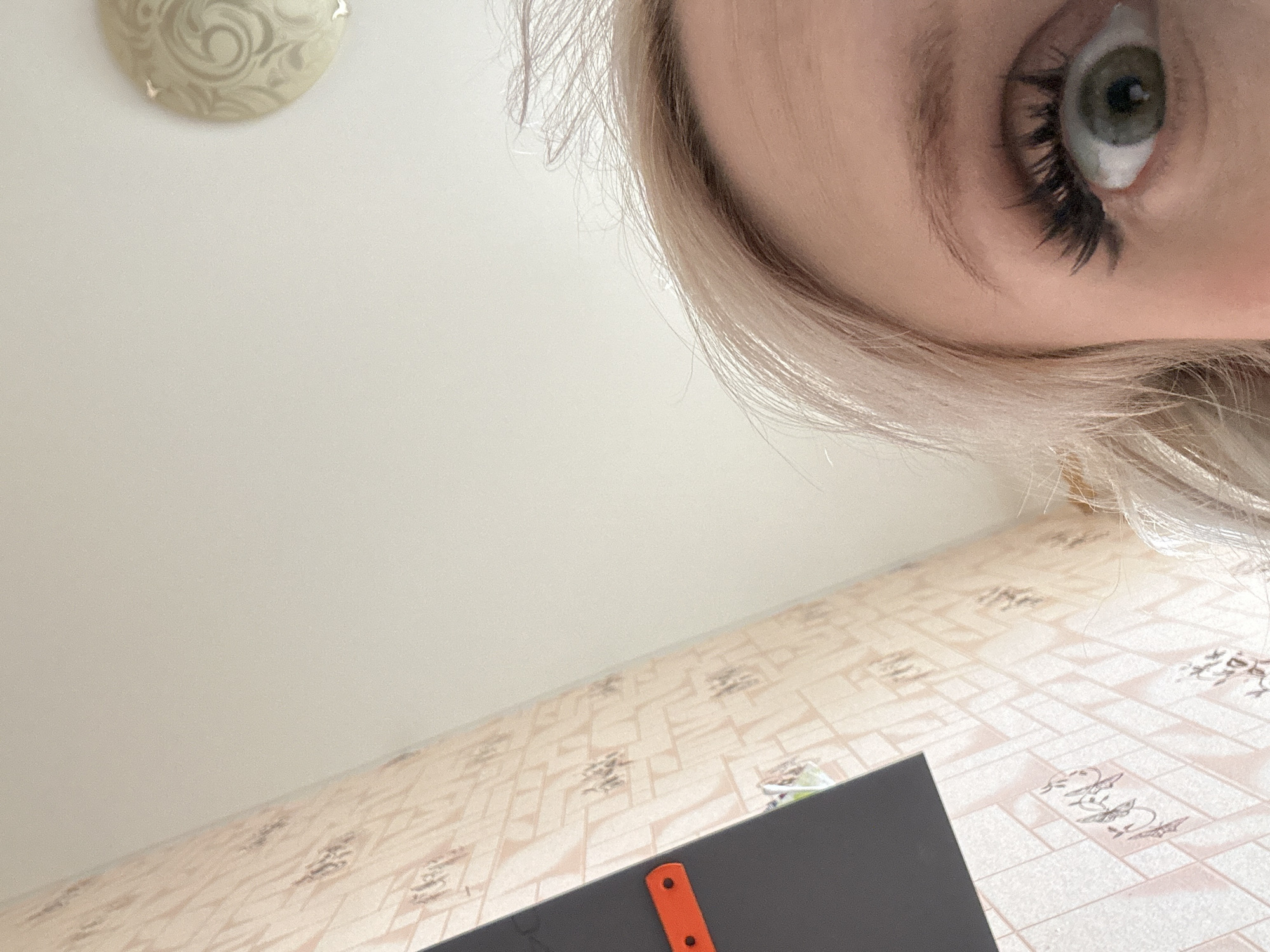 An upward-looking indoor photograph captures a partial view of a person and the surrounding room. In the top right corner, a significant portion of a person's face is visible, including a light-colored eye (possibly green or blue-grey) with prominent eyelashes, a lightly arched eyebrow, and a section of blonde or light-colored hair framing the face. The person appears to be looking directly or slightly downwards towards the camera.
The ceiling, which occupies the upper left and central part of the image, is plain white or light cream. A round, decorative light fixture or ceiling medallion with swirly patterns is partially visible in the top left corner. Below the ceiling, a wall or another surface, viewed from an upward angle, is covered with patterned wallpaper or tiles. This pattern features geometric shapes like squares and rectangles, interspersed with darker, abstract motifs that could be stylized figures or symbols.
In the bottom right corner, a dark, flat object, possibly a book, tablet, or folder, is partially visible. An orange plastic item with two circular holes is attached to or resting on this dark object. The overall lighting suggests an indoor setting during daytime, with soft, diffused illumination. There is no visible text in the image. The photo provides a glimpse into a domestic setting in Stavropol, Russia.