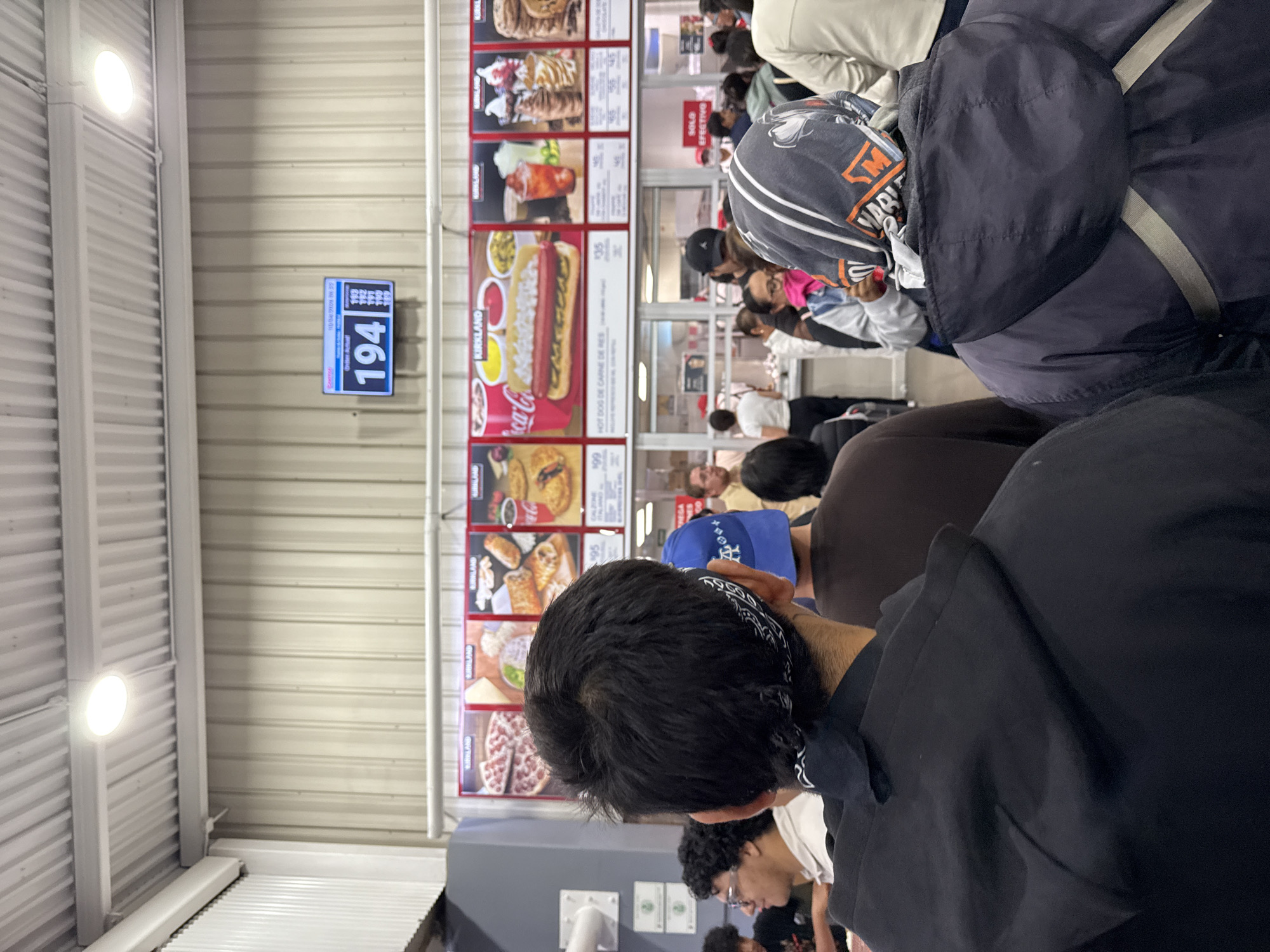 The image shows a scene from what appears to be a Costco food court. A crowd of people is gathered near the food menu board, with a digital display showing order number 194. The menu board features a variety of food items, including hot dogs, pizza, and desserts, with prices in Mexican pesos. The scene suggests a crowded and bustling environment, likely during a busy time at the store. No location in Puebla, Mexico can be identified.