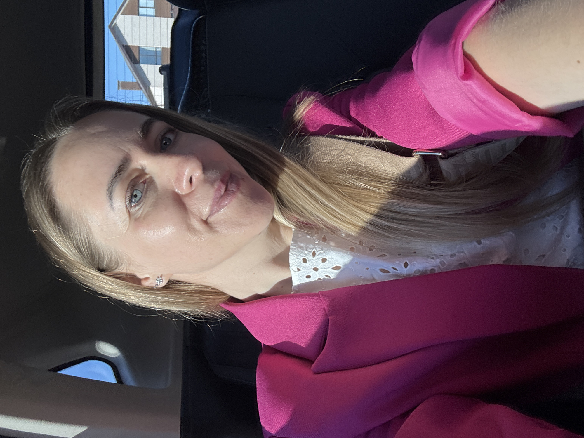 A woman with blonde hair and blue eyes is sitting in a car, possibly in Chaikovsky, Russia, with a building visible through the window. She is wearing a white, eyelet-trimmed top under a bright pink blazer. The image is taken from a close-up, slightly angled perspective. The light is bright. She has a silver colored earring on her left ear.