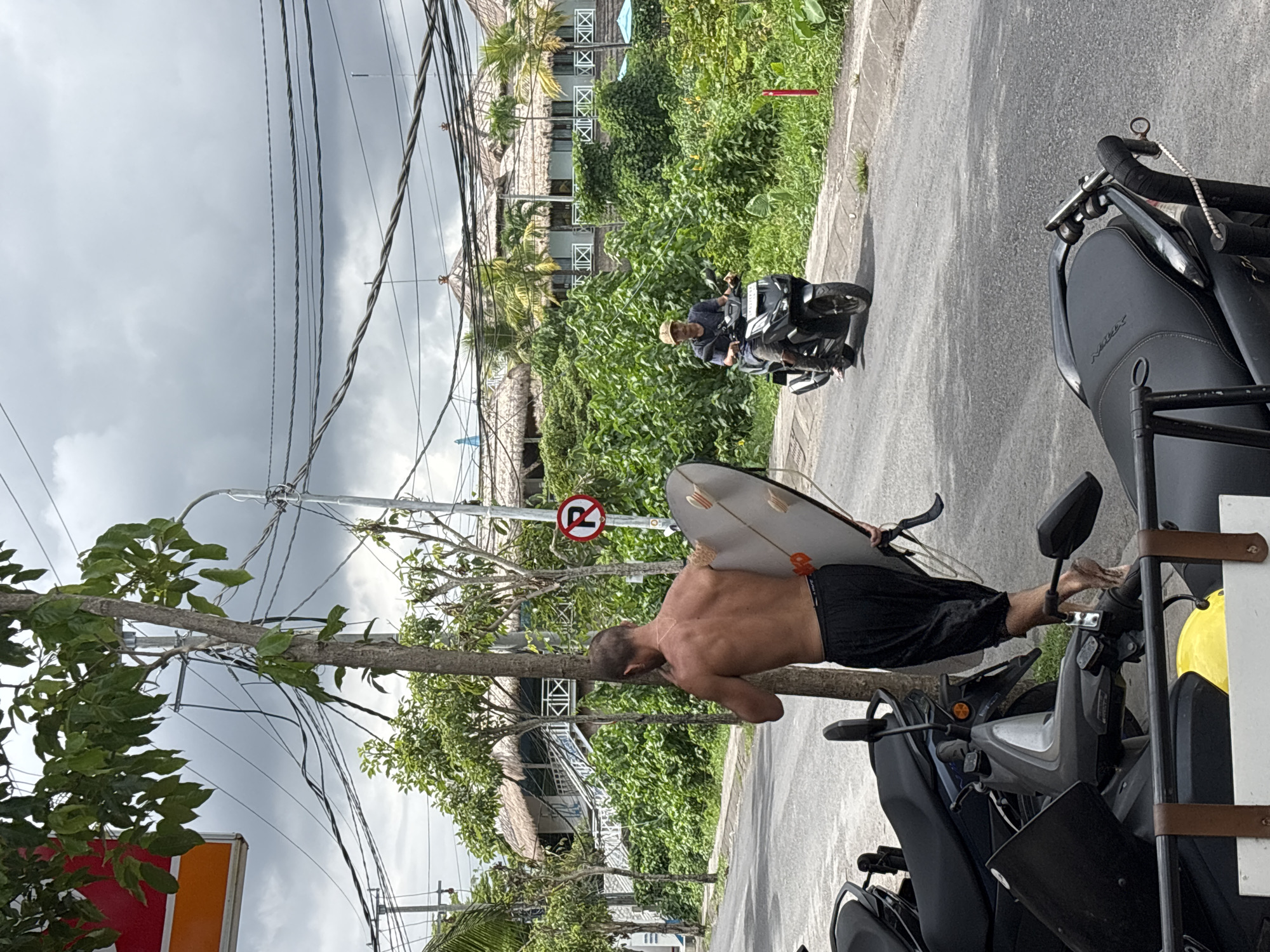 On a paved street in Mengwi, Indonesia, a shirtless man in dark shorts stands beside parked black scooters. He rests his head on a tree trunk near a "no parking" sign, a surfboard under his arm. Another man on a scooter is further down the street, against a backdrop of lush greenery, thatched roofs, and buildings with white railings. Overhanging are utility poles and wires against a cloudy sky. A partial commercial sign is visible in the lower left.