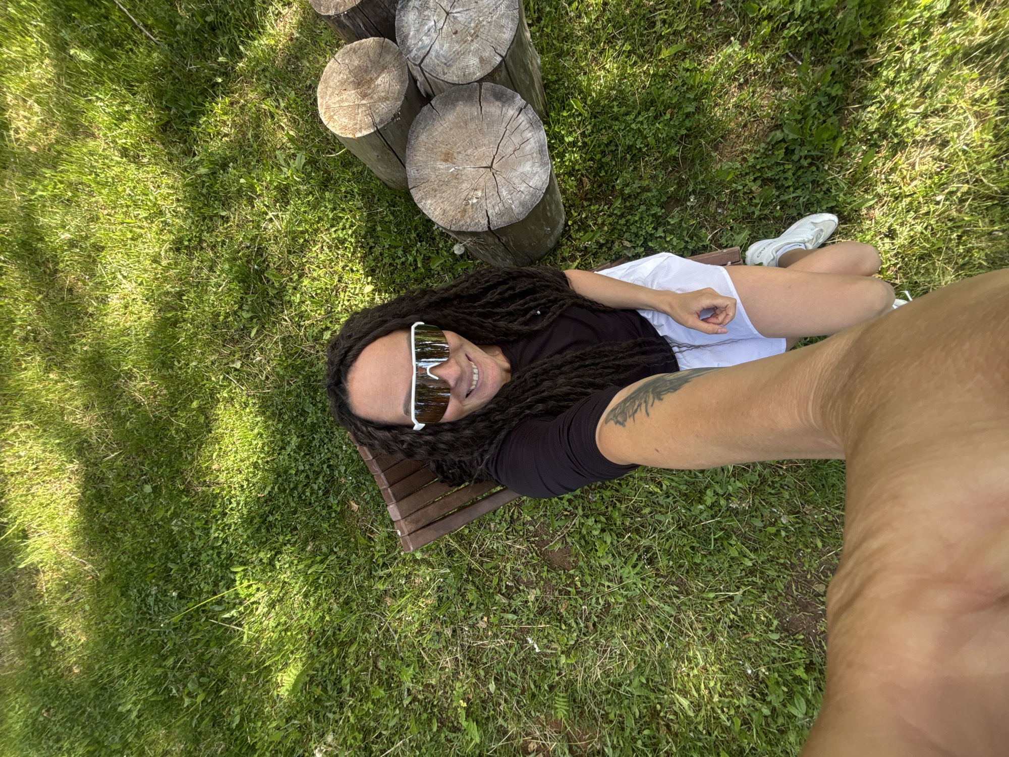 A high-angle shot captures a person lying on a small wooden bench in a grassy area, likely a park. The person has dark dreadlocks and wears white sunglasses, a black shirt, and white shorts. Their left arm, extended towards the camera, features a tattoo. To the left of the bench are three stacked wooden logs. A pair of white sneakers are visible near the person's legs. The grassy area appears to be well-maintained. Based on contextual information, the location is potentially in Chaikovsky, Russia.