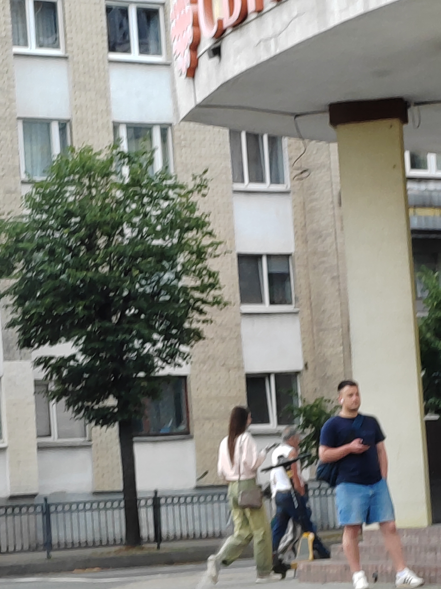 An outdoor, ground-level, medium shot shows a street scene with a building and several people. In the background, a light-colored, multi-story building with many windows and a curved awning is visible. In the foreground, a young woman in a light pink shirt and olive green pants walks from left to right, carrying a brown crossbody bag. To her right, a man wearing a dark blue t-shirt and light blue denim shorts stands looking to his right, holding a phone in his right hand. He has a blue bag slung over his left shoulder. Behind him, partially obscured by the man, is another person, possibly a man in a white shirt and dark pants, carrying a dark object. A tree with dark green foliage is positioned to the left of the woman, between her and the building. A metal fence runs along the bottom of the frame. The scene appears to be in Brest, Belarus, based on the context provided.